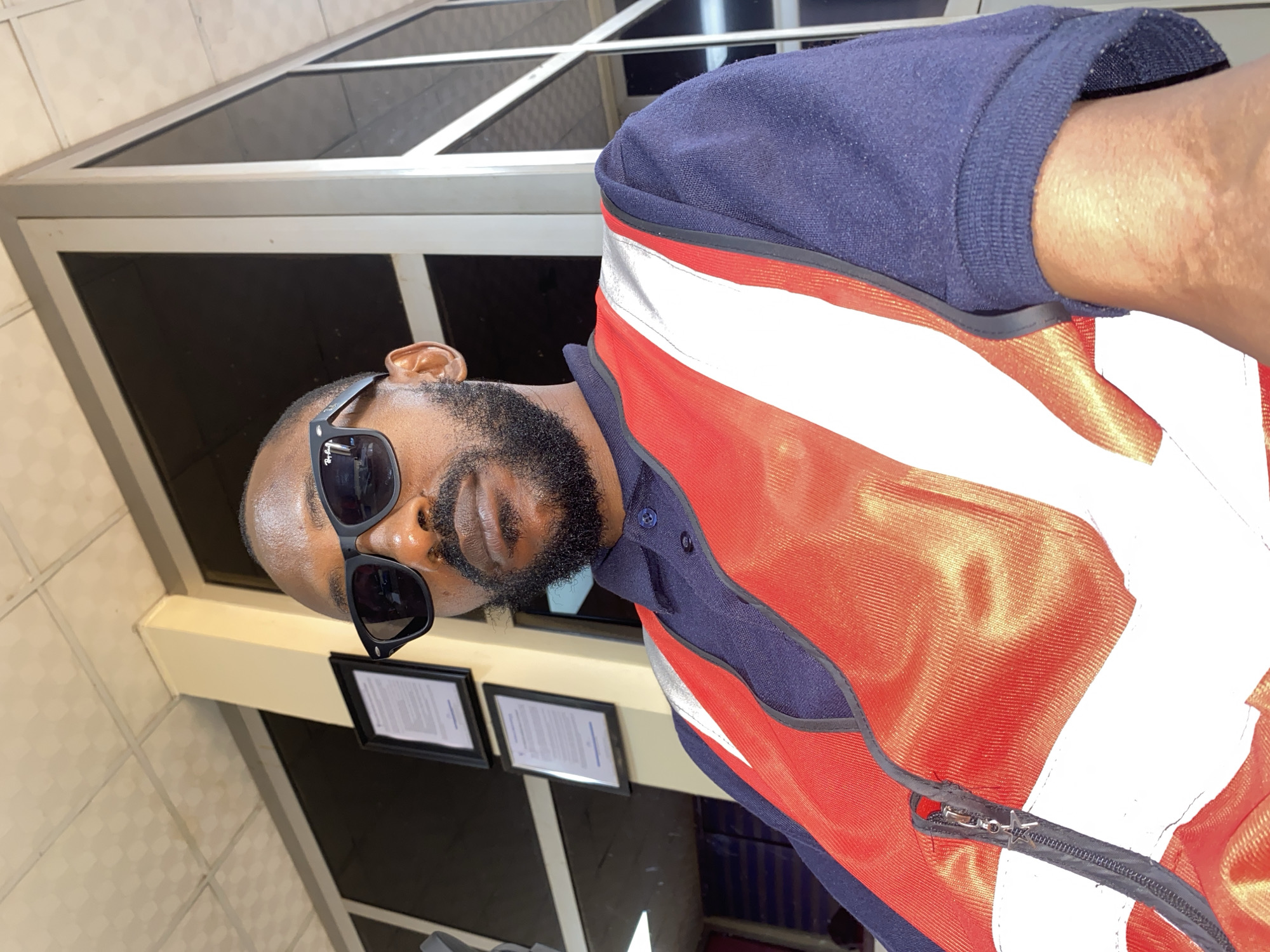 The image captures a close-up portrait of a Black man, presented at an oblique angle as if taken from slightly below. He has a well-groomed dark beard and mustache, and his head is mostly shaved on the sides. He is wearing black sunglasses, which feature the visible "Ray-Ban" brand name on the arm, obscuring his eyes. His expression is neutral and composed.
He is dressed in a dark blue long-sleeved collared shirt, layered with a prominent bright orange or red high-visibility vest or jacket. This outer garment is designed with broad white reflective stripes across the chest and back, and a dark zipper runs down its front.
The background indicates an indoor or semi-indoor setting. Behind the man are large, dark-framed windows or glass panels reflecting ambient light. Below these, a light-colored wall, possibly tiled, is visible, featuring two framed documents with text that is not legible. A light-colored architectural beam or ceiling structure is also present. The scene suggests a professional or public building environment, and the man appears to be in a work uniform. The image is likely a selfie or a casual portrait.