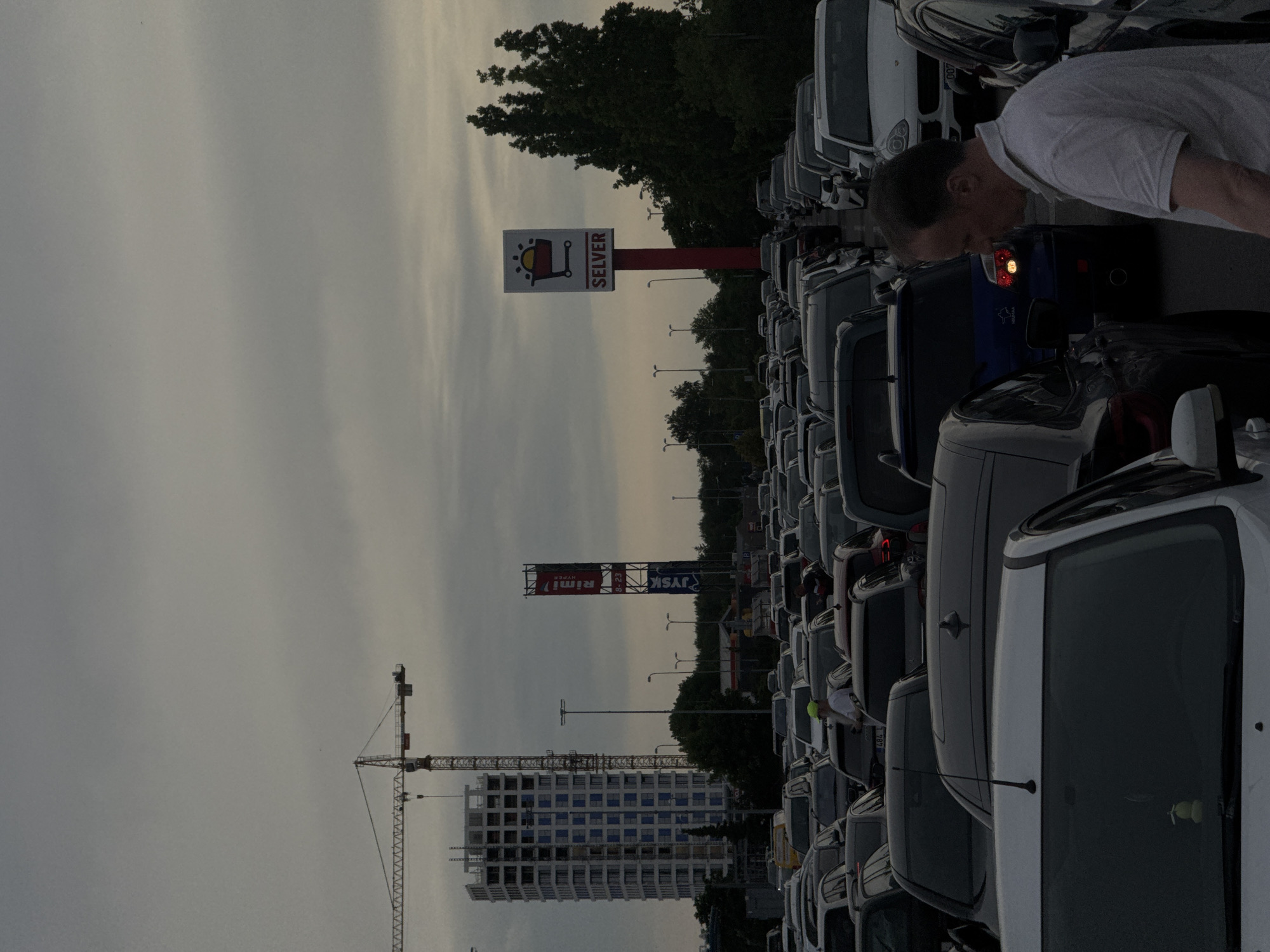 The image depicts a traffic jam in a parking lot, likely at a commercial area, under an overcast sky. In the background, a tall, unfinished building under construction is visible, with a construction crane positioned to its left. A red and white sign for "SEVER" with a shopping cart icon is prominent on a pole. Further to the left, a tall red and white sign with "Rimi" and "JYSK" branding is also visible. The parking lot is filled with numerous cars, and a man in a light-colored polo shirt is walking among them, looking towards his right. The scene suggests a busy commercial district with ongoing development.