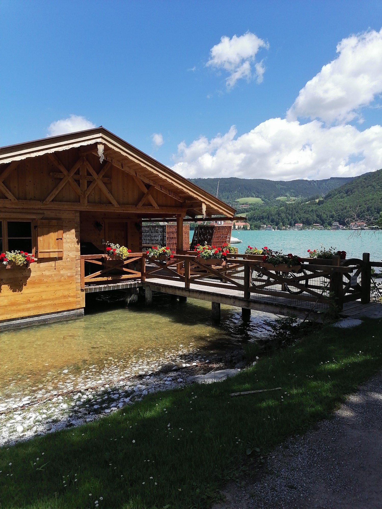This image shows a wooden boathouse on the edge of Lake Tegern in Rottach-Egern, Germany. The boathouse is constructed with exposed wooden beams and features a sloped roof with dark trim. Flower boxes filled with red and yellow flowers adorn the windowsills and the railing of the attached walkway. The water is clear, revealing stones on the lakebed. A wooden walkway leads from the boathouse to the shore, where lush green grass and a gravel path are visible. In the background, verdant hills rise, dotted with buildings and trees, under a bright blue sky with scattered white clouds. The scene depicts a serene lakeside environment on a sunny day.