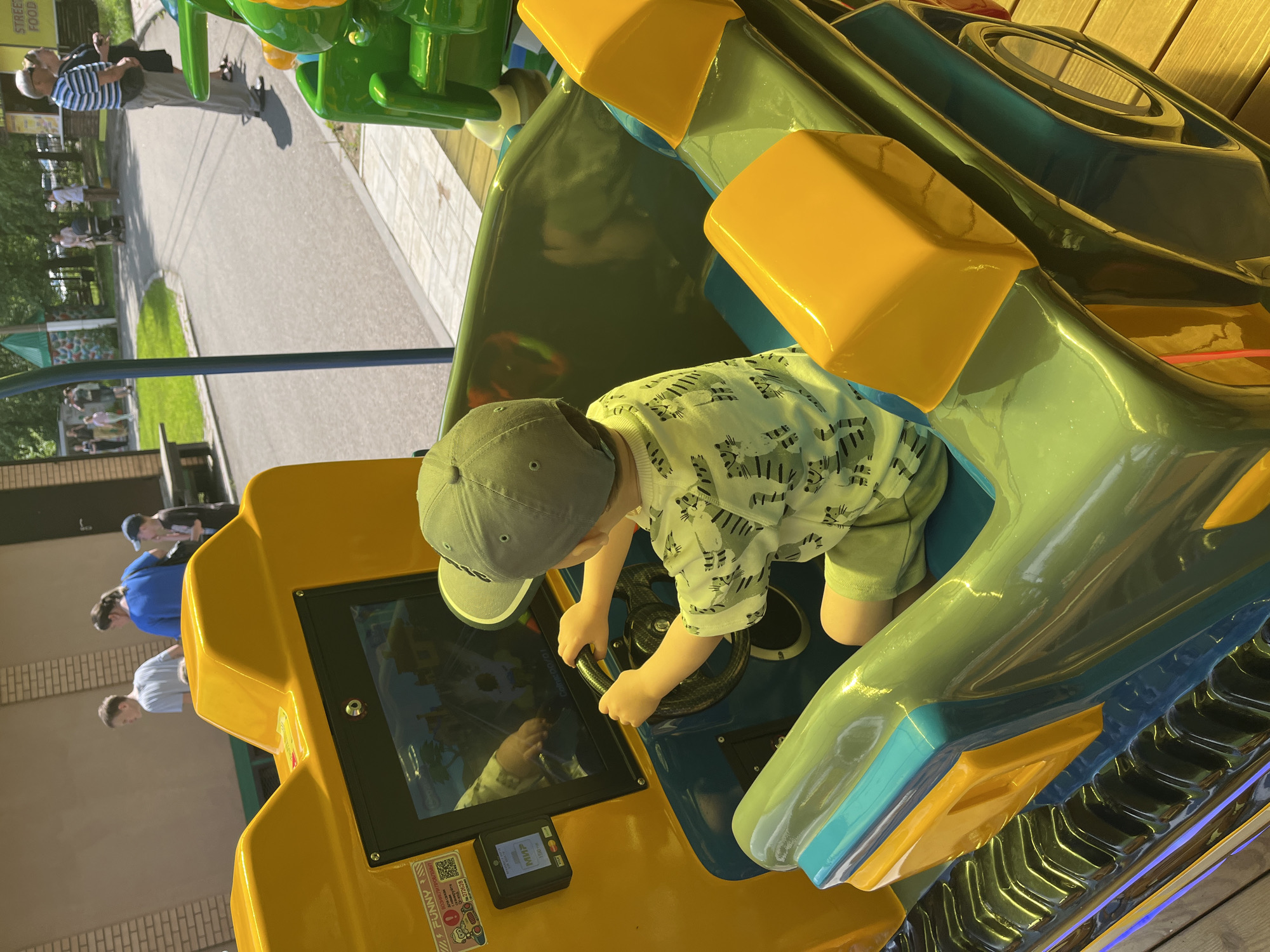 An indoor amusement park ride has a young boy wearing a grey baseball cap and a white t-shirt with a black tiger pattern, playing a video game. The boy is seated in a green and yellow vehicle with a steering wheel and a screen displaying a cartoonish landscape. He is holding the steering wheel with both hands, and his reflection is visible on the screen. The background shows a playground with other children and adults, and a paved pathway. The location is identified as an amusement park in Chaikovsky, Russia.