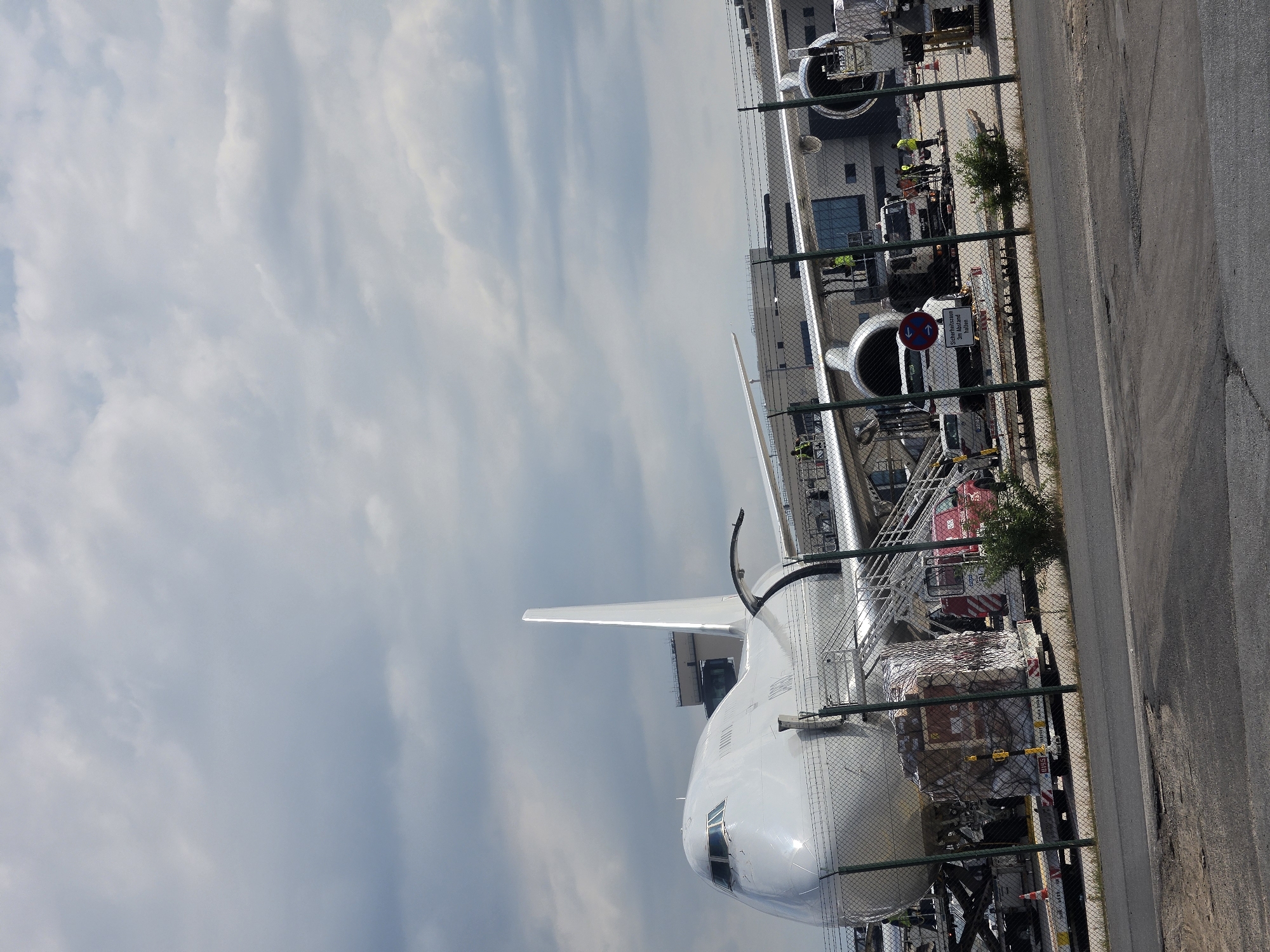 An aerial view shows a large, white cargo airplane with its nose facing right, parked on an airport tarmac. The aircraft has four engines under its wings, and a tall, vertical stabilizer at the rear. Multiple cargo containers are visible on dollies near the plane, some being loaded or unloaded by airport personnel. Other ground vehicles, including trucks and what appears to be a small car, are also present. In the background, a long, modern airport building with large windows is visible. The scene is framed by a wire fence. The sky above is partly cloudy. The image was taken at Frankfurt Airport (FRA) in Frankfurt, Germany.