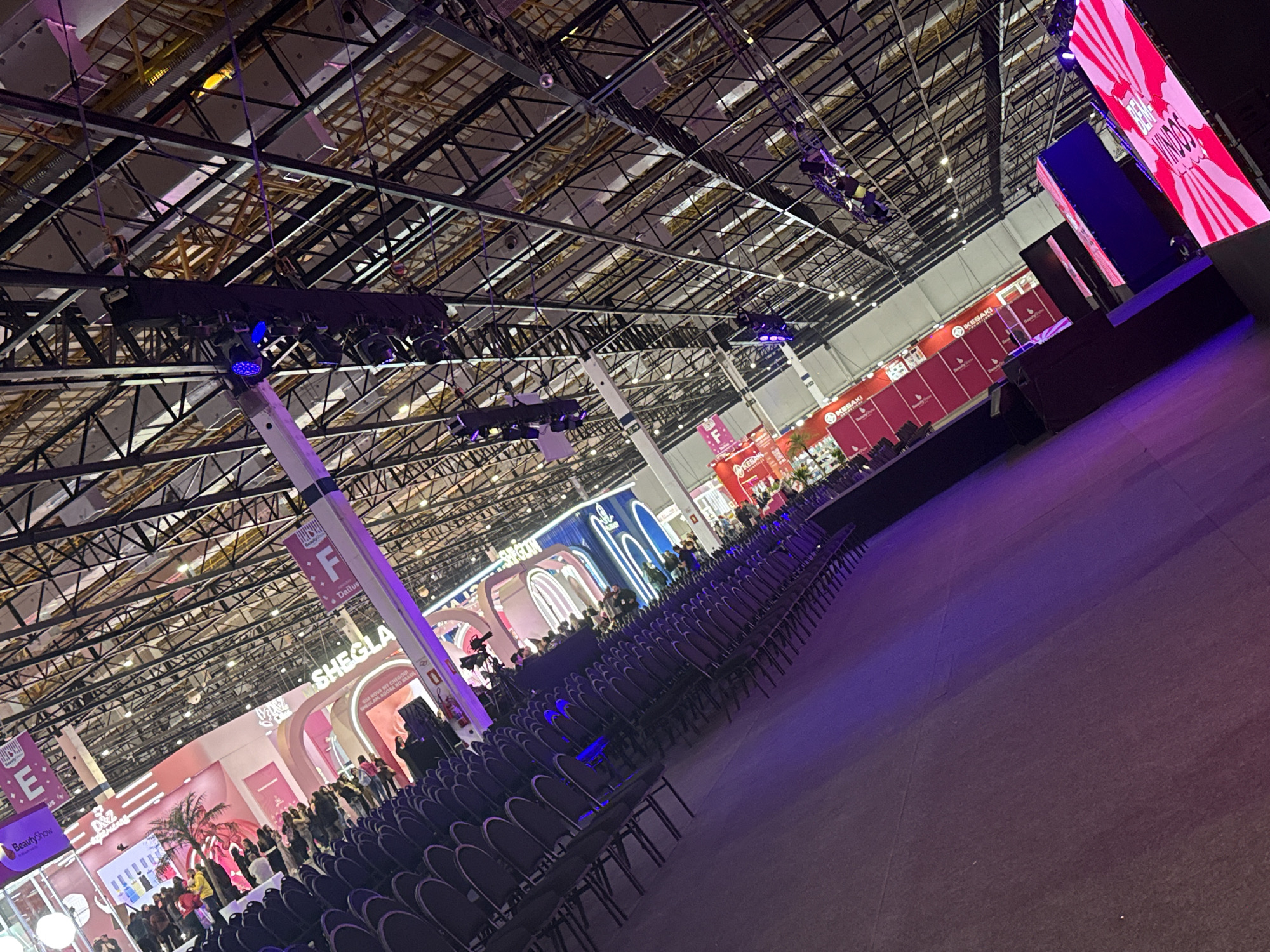The image shows an indoor event space, possibly a convention or trade show, in the city of São Paulo, Brazil. The venue has a high ceiling with exposed metal beams and rigging. A stage is set up at the far end, with a large video screen displaying text in Portuguese. Rows of empty chairs fill the foreground, leading towards the stage. Several booths and exhibits are visible in the background, featuring branding for various beauty and cosmetics companies like Sheglam, BeautyShow, and Kesaki. People are present in the background, primarily around the booths and exhibits. The lighting is a mix of natural light from the ceiling and artificial lighting, with some areas illuminated in purple.