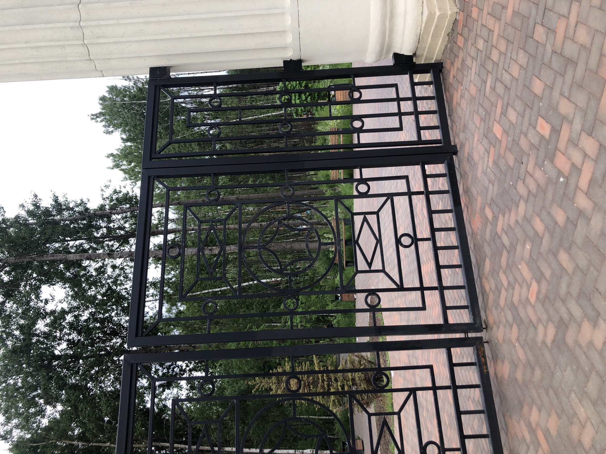 A tall, fluted, cream-colored column is to the right of the image. To the left of the column is a closed, black, wrought-iron gate that is divided into two sections. The gate has geometric patterns, including squares, rectangles, and circles. Behind the gate is a park area with trees, a small evergreen tree, and a wooden bench. The ground in front of the gate is paved with interlocking bricks in a herringbone pattern.