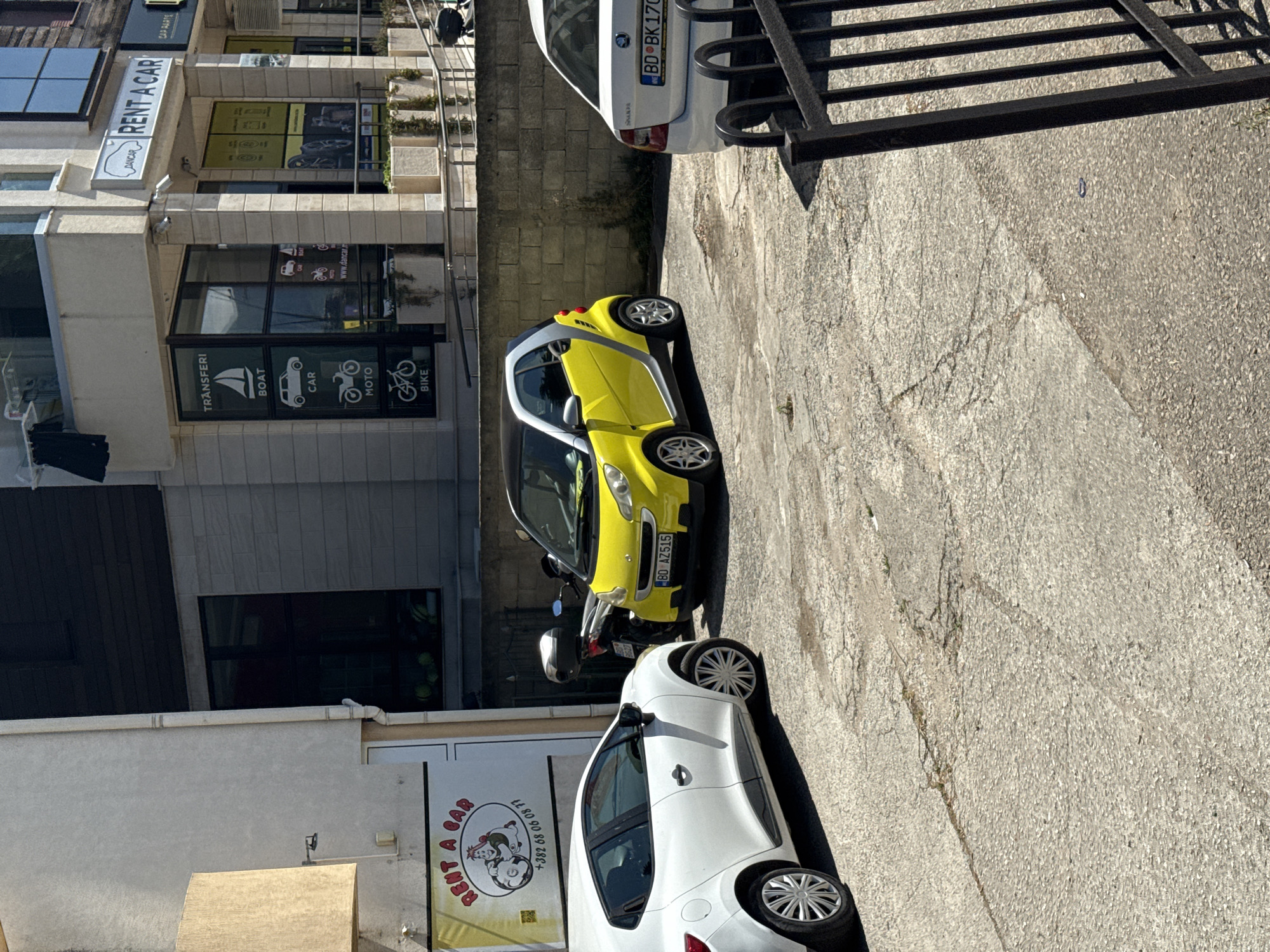 This is a medium-shot, eye-level, outdoor, daytime photograph of a rental car agency in Budva, Montenegro. The scene is dominated by two parked cars: a white hatchback on the left and a small yellow car on the right. A black motorcycle is parked behind the yellow car. The rental agency building is visible in the background, featuring large glass windows with various rental advertisements including transfers, boats, cars, motos, and bikes. One sign prominently reads "DANCAR RENT A CAR". The ground is paved with cracked asphalt. The overall lighting suggests a bright, sunny day.