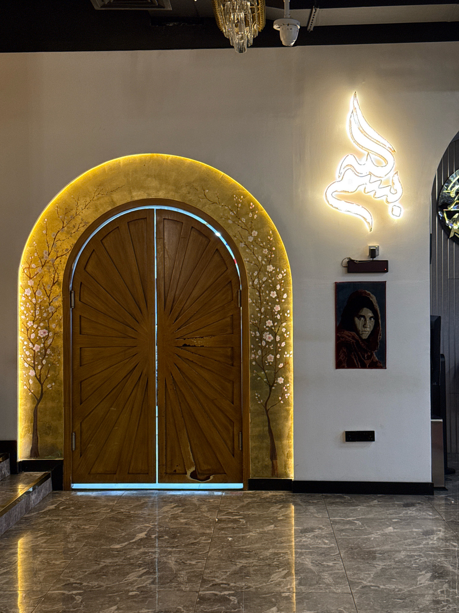 This image shows an arched wooden door in Deira, United Arab Emirates. The door is adorned with a decorative pattern resembling sunbeams. To the left of the door, there is a golden arch with painted trees and pink blossoms, illuminated by a soft yellow light. Above and to the right of the door, a neon Arabic calligraphy sign glows brightly on the white wall. Below the calligraphy, a framed portrait of a young girl wearing a headscarf is displayed. A dark power adapter is mounted on the wall above the portrait. The floor is made of polished grey marble tiles, reflecting the ambient light. To the upper left of the frame, part of a modern chandelier is visible, and a ventilation grate is present on the ceiling.