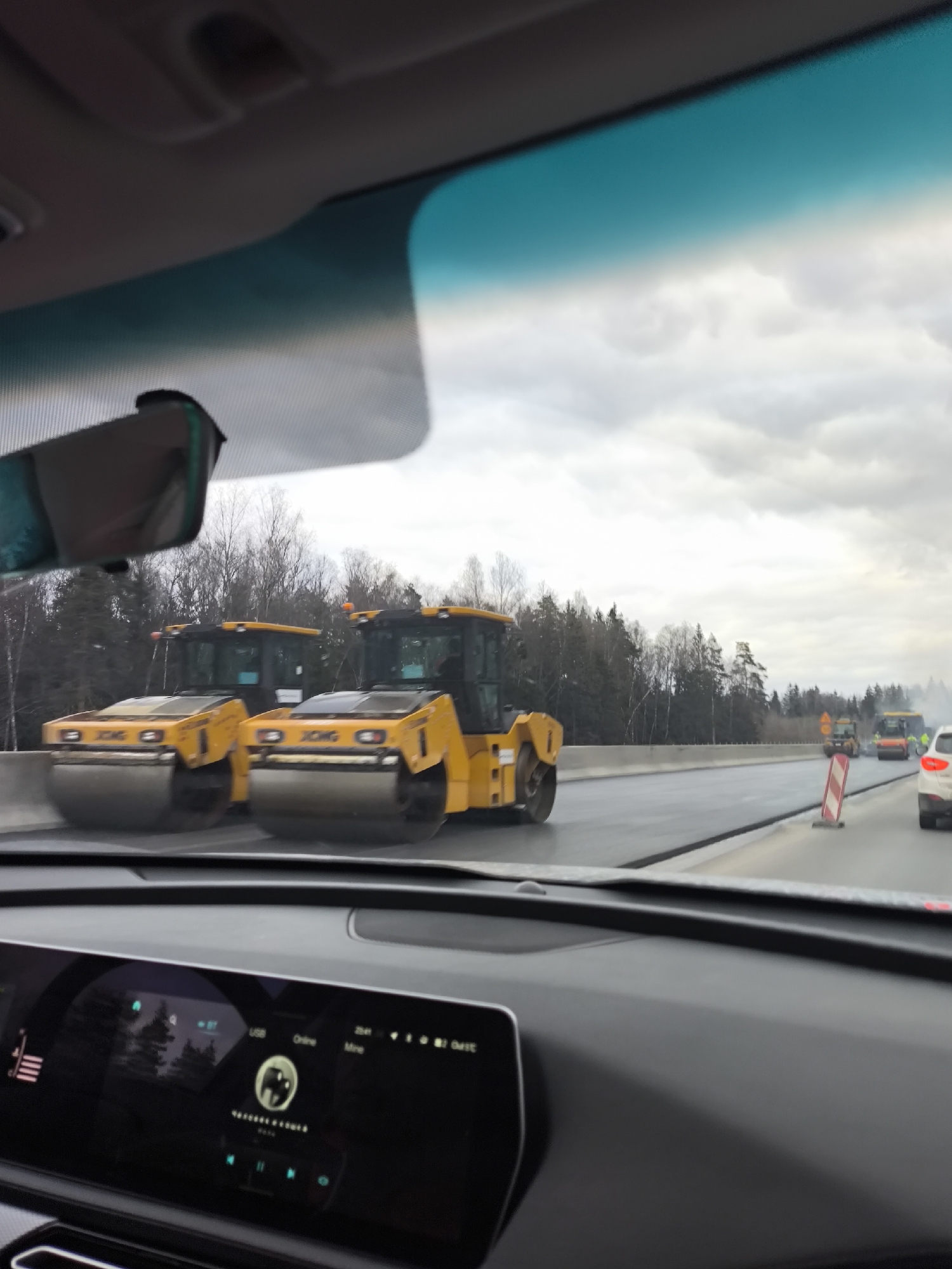 The image depicts a view from inside a car, likely on a highway or major road under construction. Two yellow XCMG road rollers are visible on the road ahead, with construction cones and possibly other roadwork equipment further in the distance. The surrounding environment appears to be a wooded area under an overcast sky. The car's dashboard is visible in the foreground, with a digital display showing car information. No specific landmarks identifying the location as Kapan', Russia, are discernible in the image.