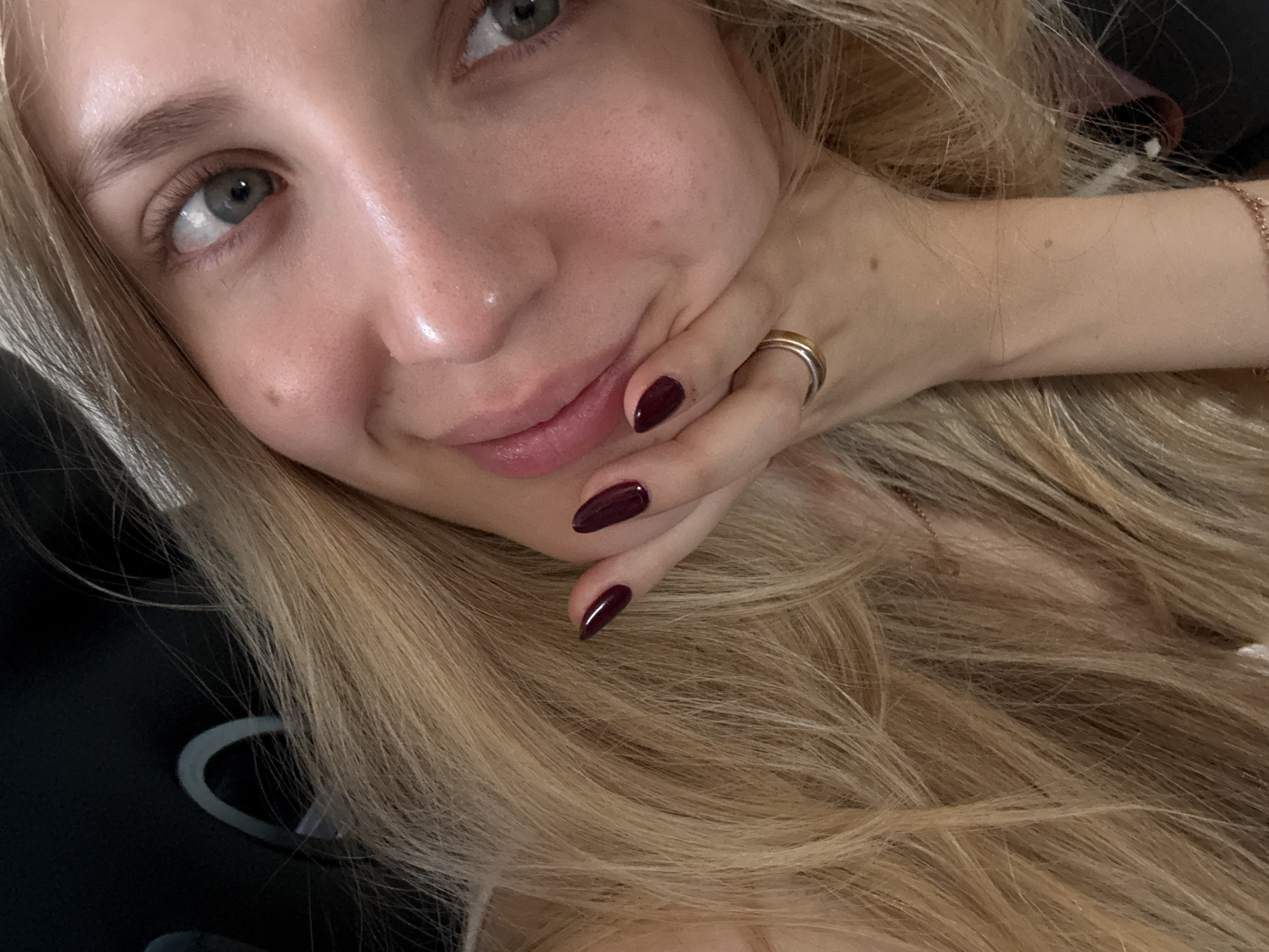 A close-up portrait captures a person with fair skin and long, wavy blonde hair. The subject, looking directly at the camera, has green/hazel eyes and a gentle, pleasant smile. Their left hand, featuring dark red/burgundy nail polish and a two-toned ring (gold and silver bands) on the ring finger, is gently resting against their chin and cheek.
Notable details include natural-looking eyebrows, prominent eyelashes, and subtly tinted lips. Several small moles or beauty marks are visible on the face and the exposed arm. The blonde hair, with lighter highlights, flows abundantly, framing the face and hand. A partial view of a delicate bracelet is visible on the wrist. The background is dark and out of focus, suggesting an indoor setting or a shallow depth of field. Vague shapes, possibly parts of clothing or an object, are indistinctly visible in the blurred background. No text is visible in the image. The time of day and specific location cues are not discernible from the photograph itself.