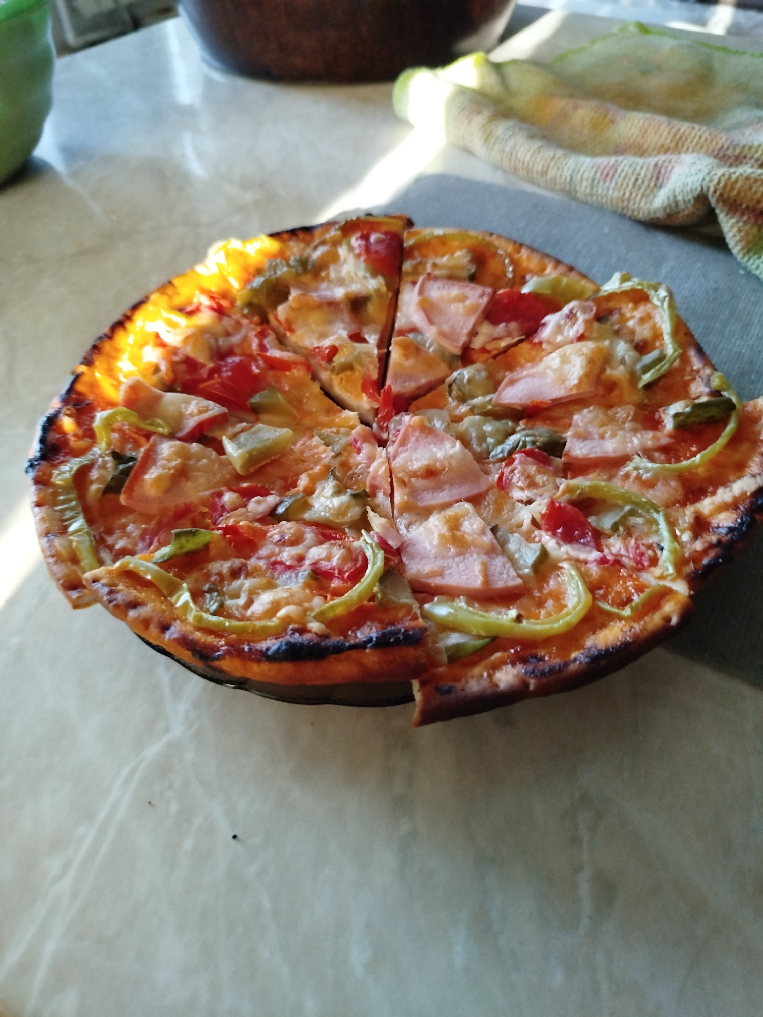 A whole, sliced pizza with ham and green peppers sits on a speckled, light-colored table. The pizza has a slightly burnt crust and is topped with melted cheese, red sauce, and visible pieces of ham and green bell peppers. In the background, a woven cloth lies on the table, and a dark bowl is partially visible. The lighting suggests it is daytime, with sunlight creating bright highlights and shadows on the scene. No individuals are present. This image was taken indoors, likely in a home setting, and there are no specific identifying features that place it in Zavodoukovsk, Russia.