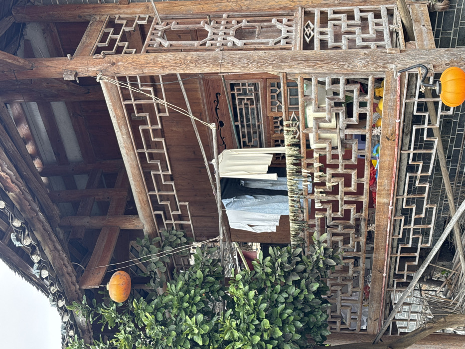 This image depicts a multi-story, traditional wooden structure in Furong, China, under an overcast sky. The building features dark wooden beams, panels, and intricate geometric lattice-work railings on its upper and lower visible levels. A dark tiled roof with exposed rafters crowns the structure.
On the upper balcony, three items of clothing—a light-colored long-sleeved shirt, a dark garment (possibly pants or a shirt), and a cream-colored long-sleeved shirt—are suspended from a clothesline, which is strung between two wooden supports with visible ropes. Directly below the clothesline, a horizontal row of light-colored, stringy, dried items, resembling herbs or food strips, is draped over the balcony's lower railing. Through the upper balcony's ornate railing, various colorful objects and some yellow/orange items are visible on the floor, along with a small white figure resembling a cat or statue.
Two orange, pumpkin-shaped lanterns are present: one hangs from a tree branch on the far left, near the building's roofline, and another hangs from a black hook attached to a vertical wooden pillar on the lower right, beneath the upper balcony. A leafy green tree occupies the left side of the frame, partially obscuring the building's upper section. The building's base features a dark brick wall section.