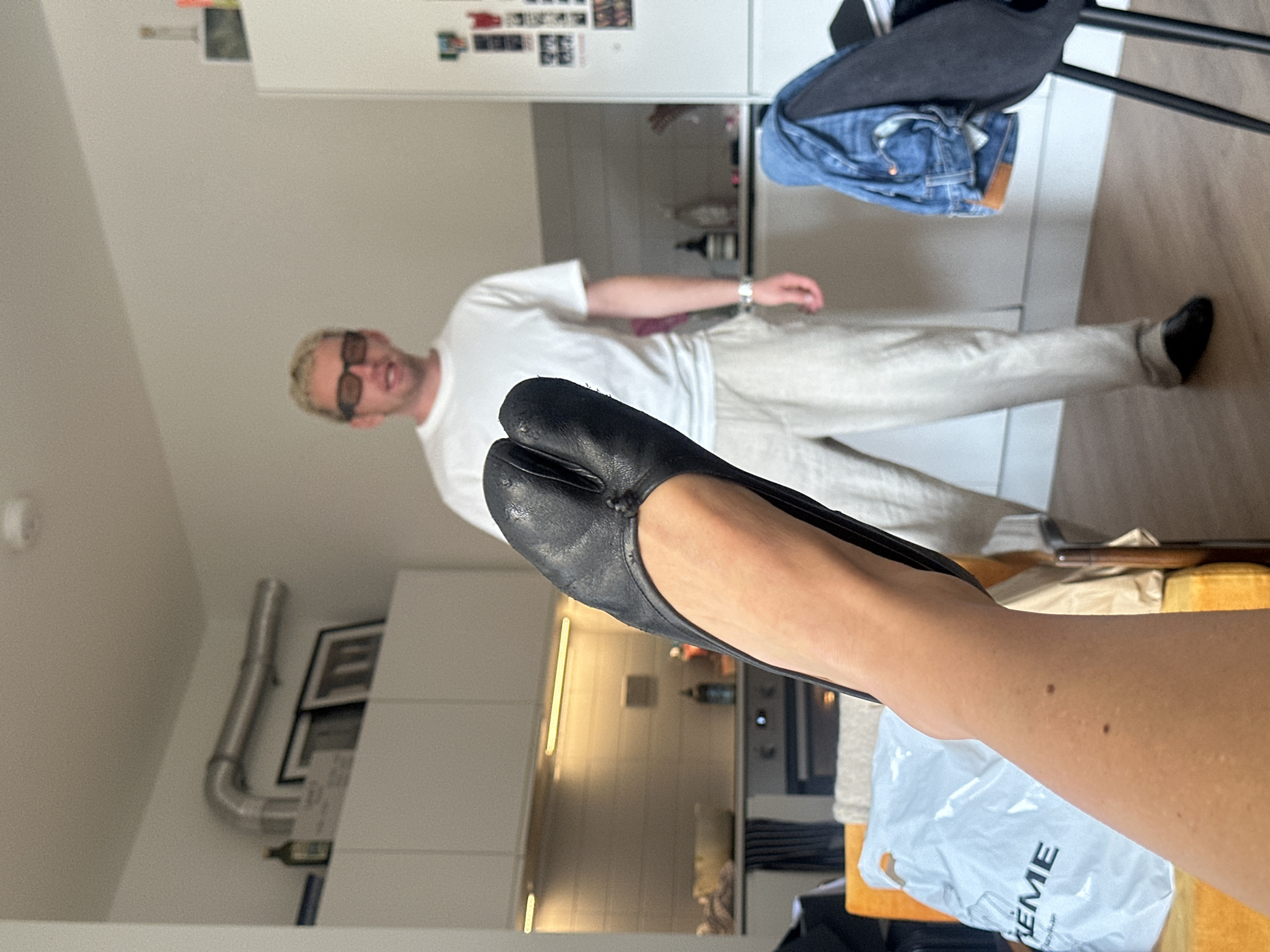 In a kitchen and living area of a London residence, a person's leg and foot, wearing a black split-toe shoe, are prominently featured in the foreground. The shoe is black leather and appears to be a flat style with a distinct split between the big toe and the other toes. Behind this, a man with blonde hair and dark-rimmed glasses stands wearing a white t-shirt and light-colored trousers. He is positioned in front of kitchen cabinets and appliances, including a refrigerator adorned with several items. To the right, a black chair is draped with a pair of blue jeans. The overall environment suggests an indoor, domestic setting.