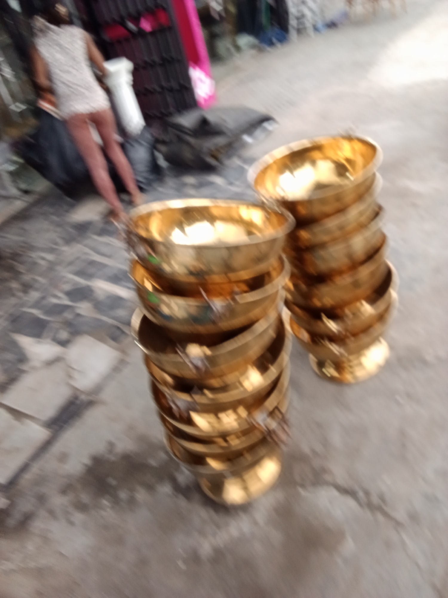 The image, taken in an urban environment, shows two stacks of shiny, golden-colored metal bowls. The stacks are positioned close to the lower right corner of the frame. To the left and further back, a person in a light-colored dress is partially visible, facing away from the camera. The foreground features a sidewalk or tiled area, and in the background, there is a structure that appears to be a storefront or building with various items displayed. The overall scene suggests an outdoor marketplace or shopping district.