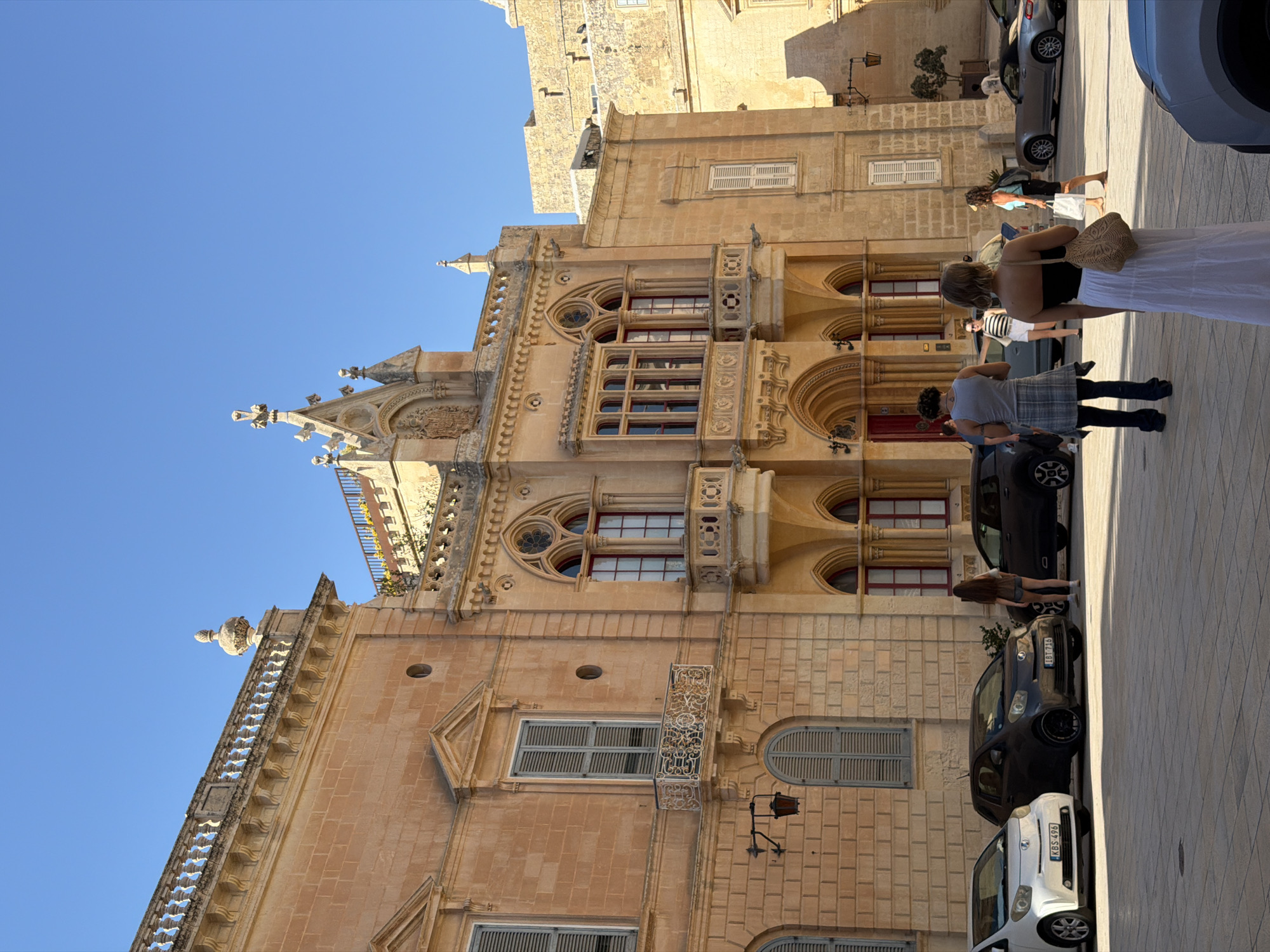 This is a daytime, eye-level shot of the city of Mdina, Malta, featuring the facade of a historic building constructed from honey-colored stone under a clear blue sky. The building exhibits ornate architectural details, including arched windows with decorative tracery, balconies with intricate metalwork, and a prominent central section with a crenellated roofline and Gothic-inspired embellishments. Several people are visible in the foreground and middle ground. To the left, three cars are parked on a paved square. In the foreground, four individuals are standing or walking. One woman, facing away from the camera, wears a white skirt and a black top, carrying a straw bag. Another person, also facing away, wears a grey tank top and a plaid skirt over dark trousers. A third person, a woman, is seen from behind wearing a striped top and white shorts. A fourth person, a woman, is partially visible in the background to the right, wearing a blue top and black skirt, carrying a white shopping bag. Further back, other figures and vehicles, including a silver convertible sports car, are present. The overall scene depicts a tourist area within the ancient walled city.