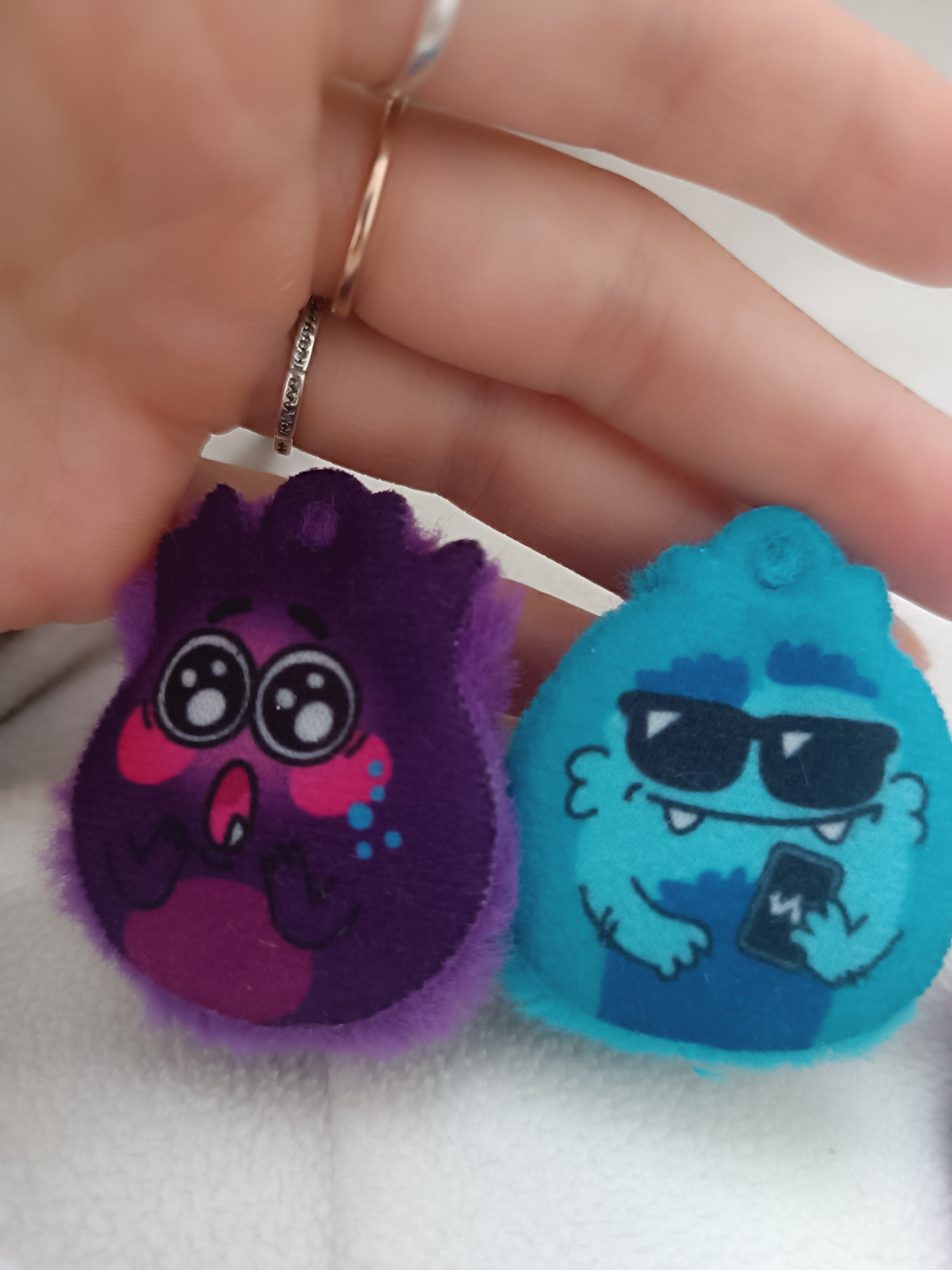 A close-up shot depicts a hand holding two plush toys. The hand is adorned with two rings on one finger, one silver with embedded designs and another plain gold band. The plush toys are cartoon monsters. The one on the left is purple with pink cheeks and large, round eyes. The monster on the right is teal and wears sunglasses, holding a smartphone in its hand. The background is a textured white surface. The location is Unknown, Unknown.