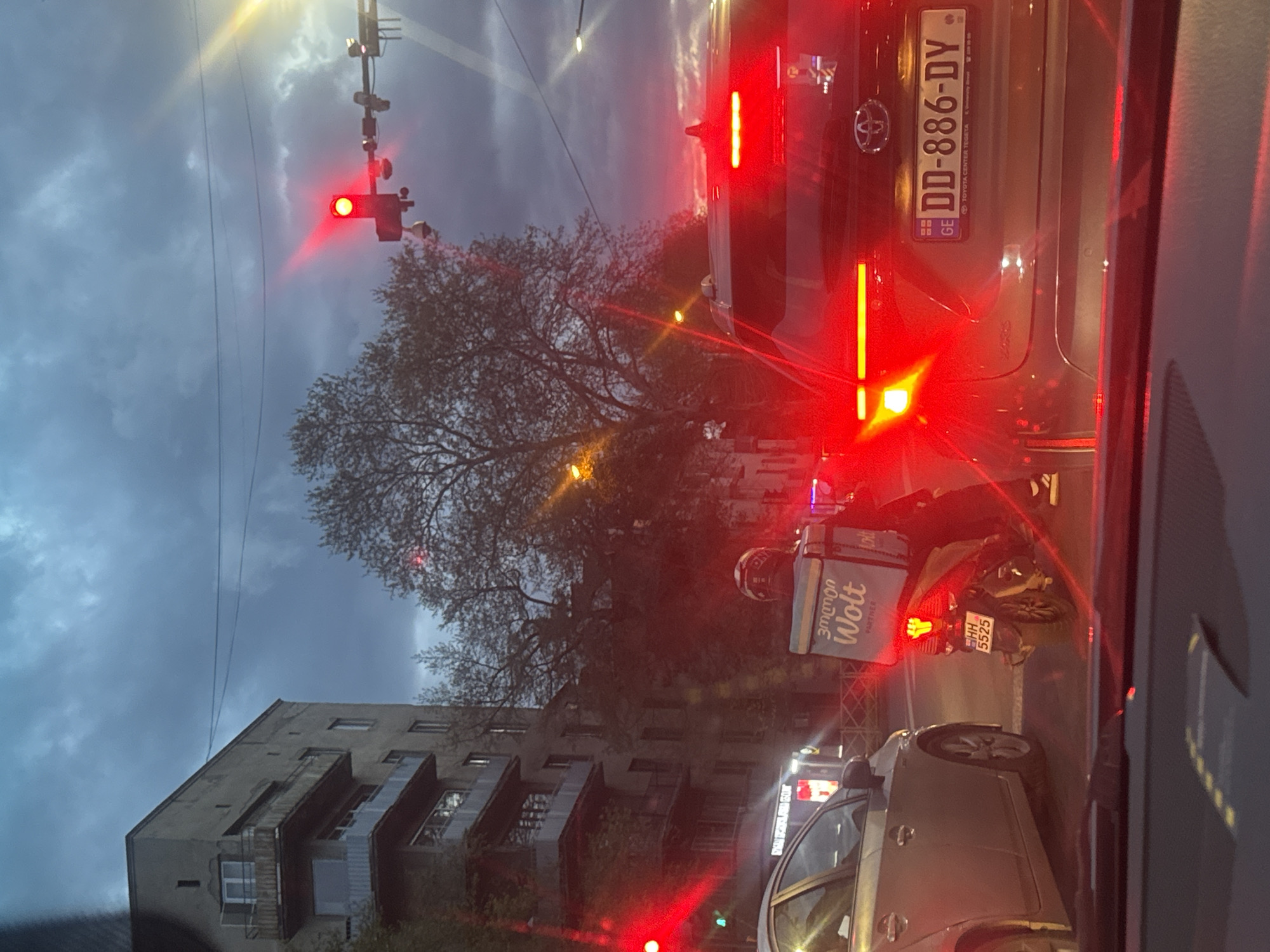 The image depicts a street scene in Tbilisi, Georgia, taken from inside a car at a red traffic light. The car in front has a license plate reading "DD-886-DY" with the "GE" country code. A Wolt delivery driver on a motorcycle is next to the car. The motorcycle's license plate reads "HH 5525." Buildings and a tree line the street, and the sky is overcast. A traffic light with a red light is visible.