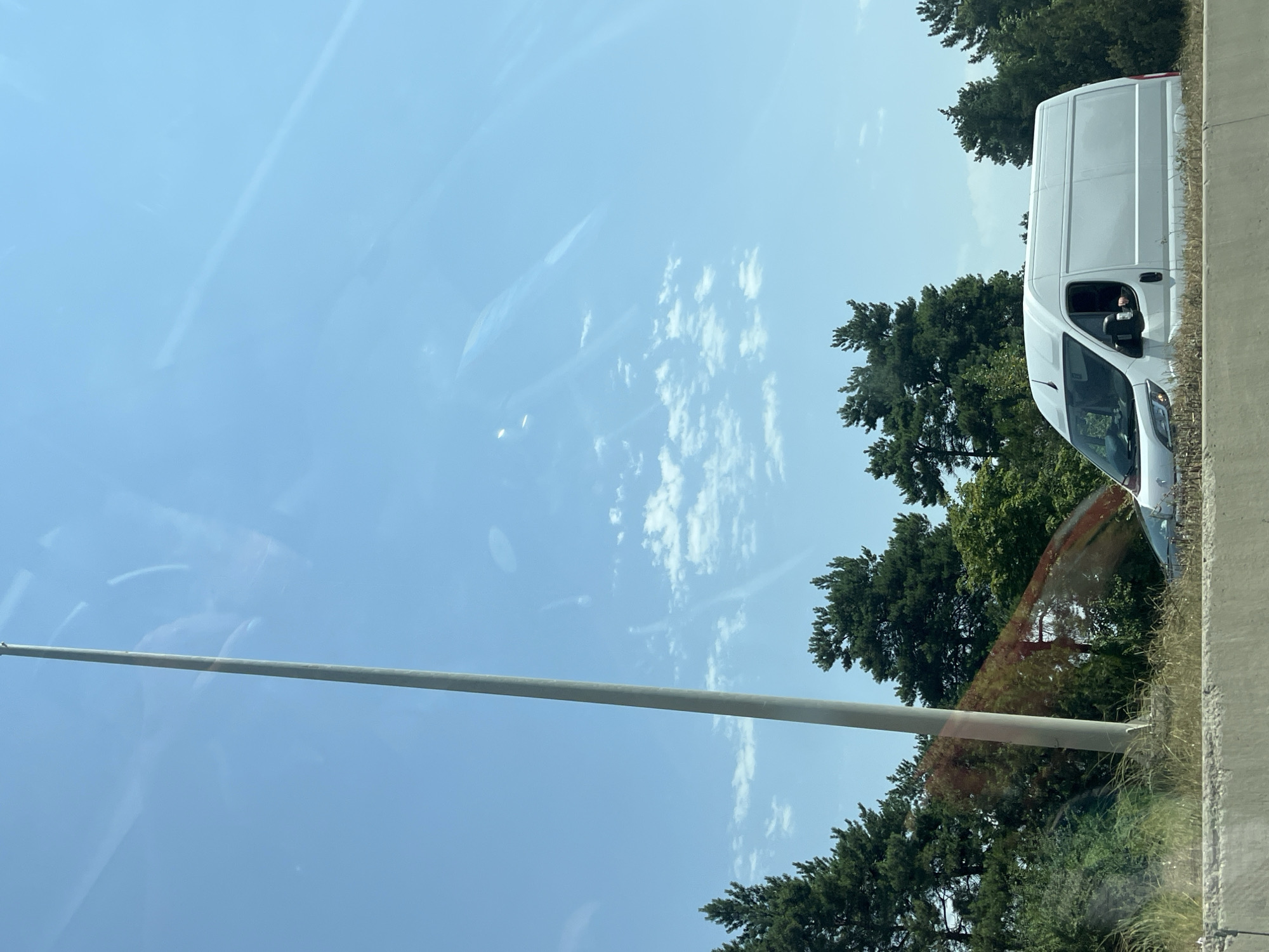An outdoor, eye-level, medium shot captures a white panel van driving on a highway under a clear blue sky with scattered white clouds. The van is in the lower right quadrant of the image, partially obscured by a concrete barrier in the foreground. Tall evergreen trees line the highway, with some deciduous trees visible in the mid-ground. A tall, slender grey pole stands vertically on the left side of the image. The scene appears to be a sunny day with good visibility. The image is taken from a vehicle, as evidenced by reflections on the windshield. The location is the city of Ruisi, Georgia.