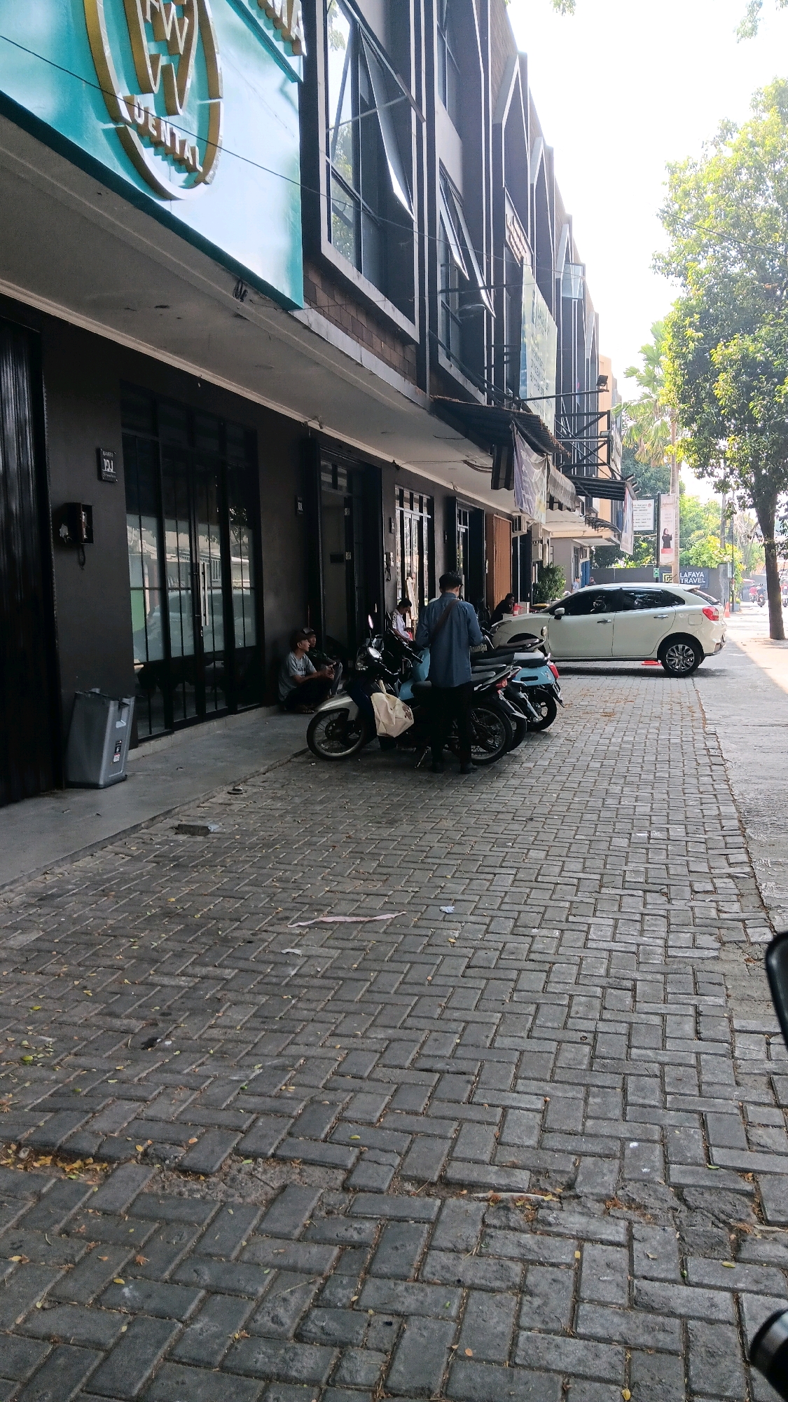 The image shows a street scene in South Jakarta, Indonesia, with a focus on a row of multi-story commercial buildings. In the foreground, a cobblestone sidewalk runs alongside the buildings. Several motorcycles are parked on the sidewalk, and a white sedan is visible on the street further down. Two individuals are sitting on the ground near the entrance of one of the buildings, appearing to be engaged in conversation. Another person stands near the motorcycles, wearing a blue shirt and carrying a bag over their shoulder. The buildings themselves have dark facades with large glass windows. A prominent sign with a gold "W" and the word "DENTAL" in teal is affixed to the upper part of one building. Trees and foliage are present, suggesting an outdoor urban environment. The lighting indicates it is daytime, possibly late morning or early afternoon, with bright sunlight casting shadows.