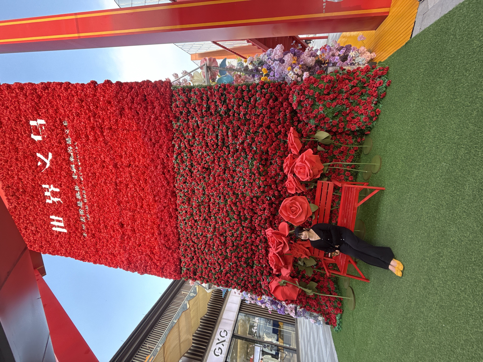 In Yiwu, China, a woman sits on a red bench in front of a large, towering floral display primarily composed of red roses. The display reaches high into the sky, with a section at the top featuring white Chinese characters. The woman, dressed in black, is angled towards the left, with her legs extended and resting on the artificial green turf that covers the ground. Her yellow shoes are visible. The bench is situated amidst oversized red rose decorations. To the right of the floral wall, a red structure with yellow accents is visible, along with a section of purple and pink flowers at the base of the rose display. In the background, a building with a store sign reading "GXG" is visible, along with a walkway and a clear blue sky. The overall scene appears to be an outdoor public space, possibly a shopping area or plaza decorated for an event.