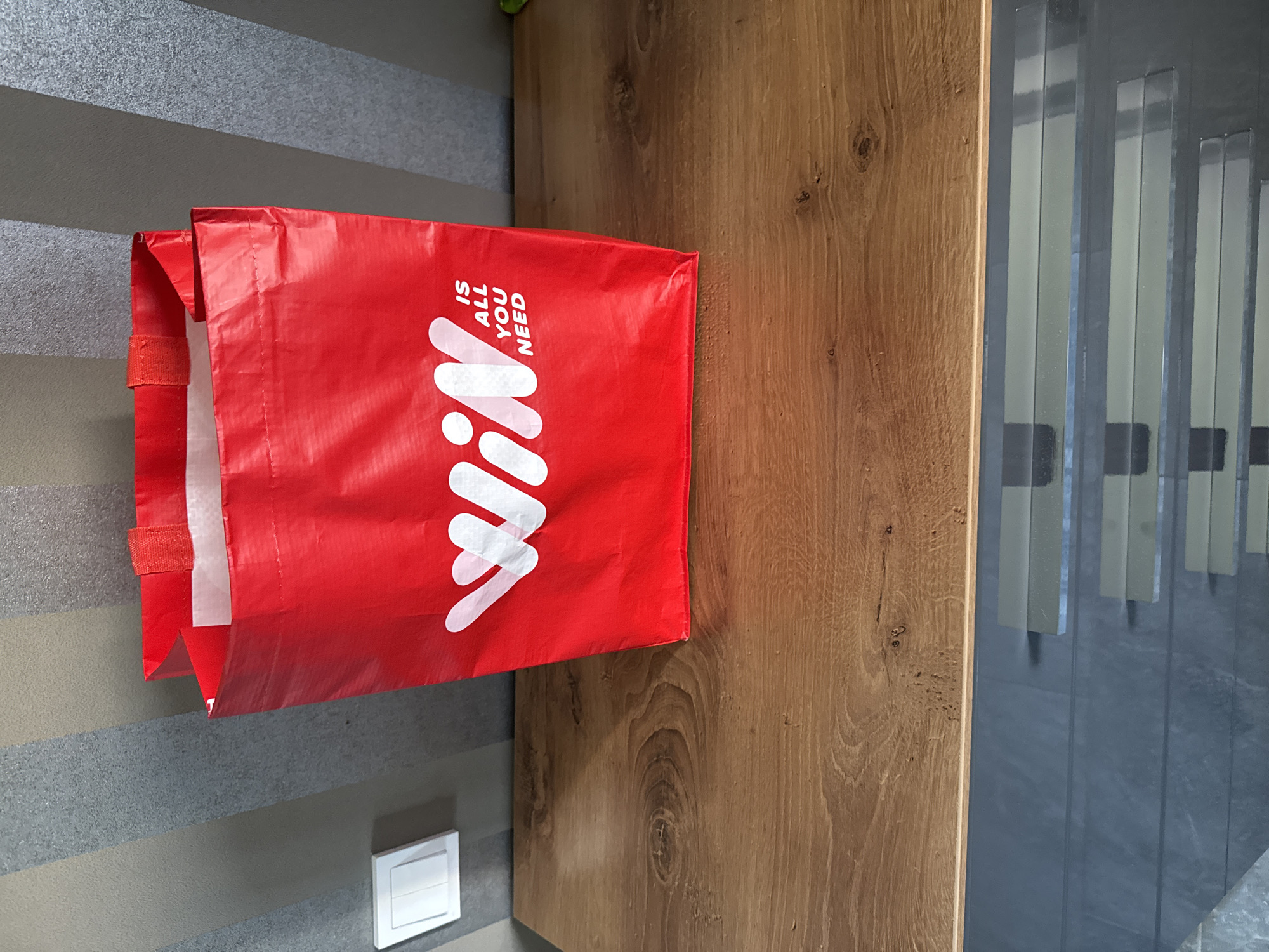 A red tote bag with white lettering that reads "WIN IS ALL YOU NEED" is placed on a wooden surface. The bag is partially open and appears to be made of a woven material. Behind the bag, a wall with vertical grey and beige stripes is visible. To the left of the bag, a white light switch is mounted on the striped wall. The wooden surface appears to be a table or shelf with visible wood grain. The overall scene is indoors, likely in a room. The location is not specified in the image itself.