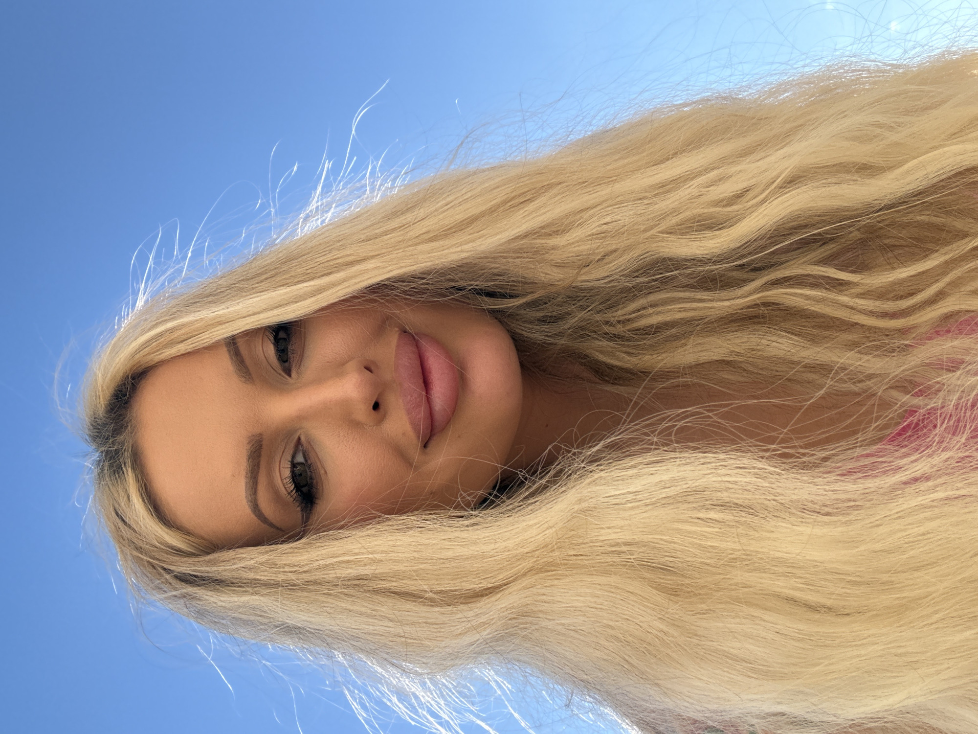 A low-angle, medium shot captures a blonde woman from the chest up, against a bright blue sky with wispy white clouds and strands of her hair blowing in the wind. The woman's face is the primary focus. She is wearing makeup, with noticeable eyeliner and lipstick. Her blonde hair is long and wavy, falling around her shoulders and framing her face. She is looking directly at the camera with a subtle smile. The background is an expanse of clear blue sky, suggesting an outdoor setting.