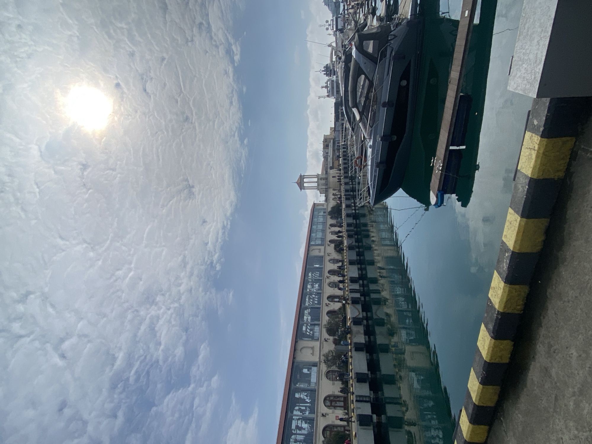 The image depicts a marina at the Sochi Seaport in Sochi, Russia, under a partly cloudy sky. The bright sun is visible in the upper right quadrant, partially obscured by widespread altocumulus clouds. The foreground consists of a concrete quay with a yellow and black striped curb on the left. The water in the marina basin is a calm, dark teal-green, exhibiting reflections of the sky and the adjacent structures.
On the left, a long, multi-story building with a light-colored facade, featuring arched ground-level openings and a continuous row of large upper-floor windows, extends across the scene. This building, part of the Sochi Seaport Passenger Terminal complex, is topped with a reddish-brown pitched roof. Numerous individuals are visible walking along the promenade in front of this building. Their actions are limited to pedestrian movement.
In the midground to the right, a dark gray or black motor yacht with a covered cockpit area and a white life preserver on its bow is prominently moored. A wooden floating dock is partially visible adjacent to this yacht. Various other boats and yachts of different sizes are moored throughout the marina basin.
In the background, behind the moored boats, a distinctive white classical rotunda structure is visible on the left, an architectural feature of the Sochi Seaport. Additional distant buildings and boats are discernible further along the waterfront. The scene is illuminated by natural daylight.