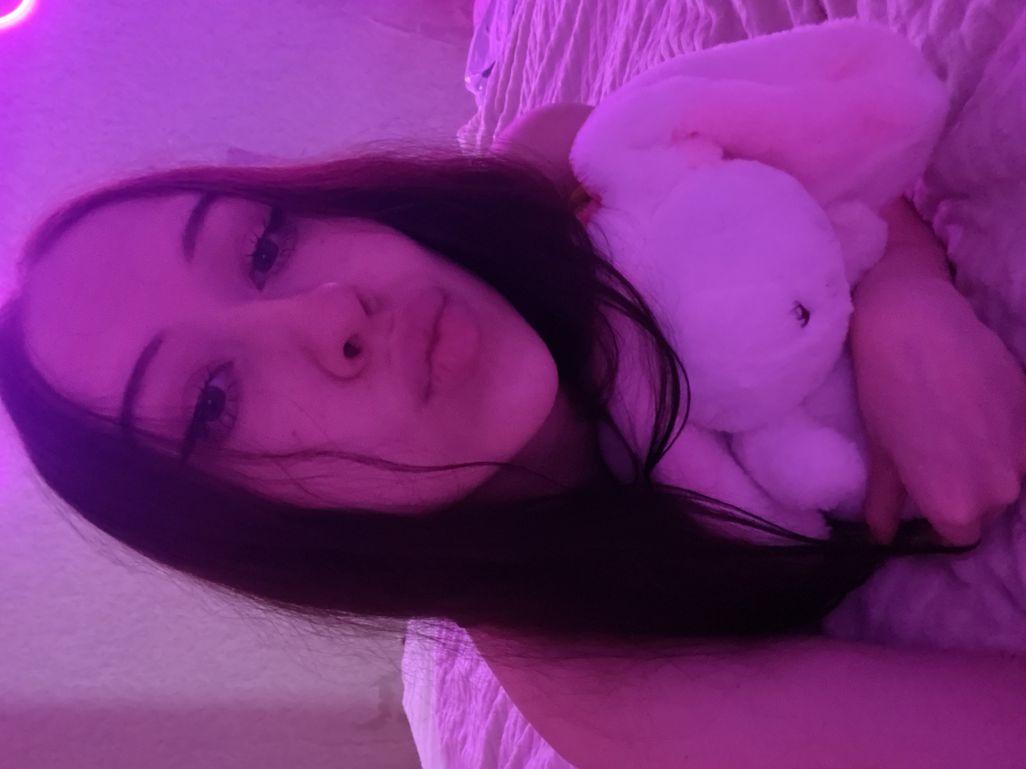The image shows a close-up selfie of a young woman with long dark hair. She is lying on a bed covered with a textured pink comforter. The room is lit with pinkish-purple lighting, possibly from a neon sign or colored bulbs. She is holding a white stuffed bunny, and her head is tilted slightly, with her lips puckered. Her eyes are looking towards the camera. The background is a pinkish wall. No specific location in Votkinsk, Russia can be determined from the image.