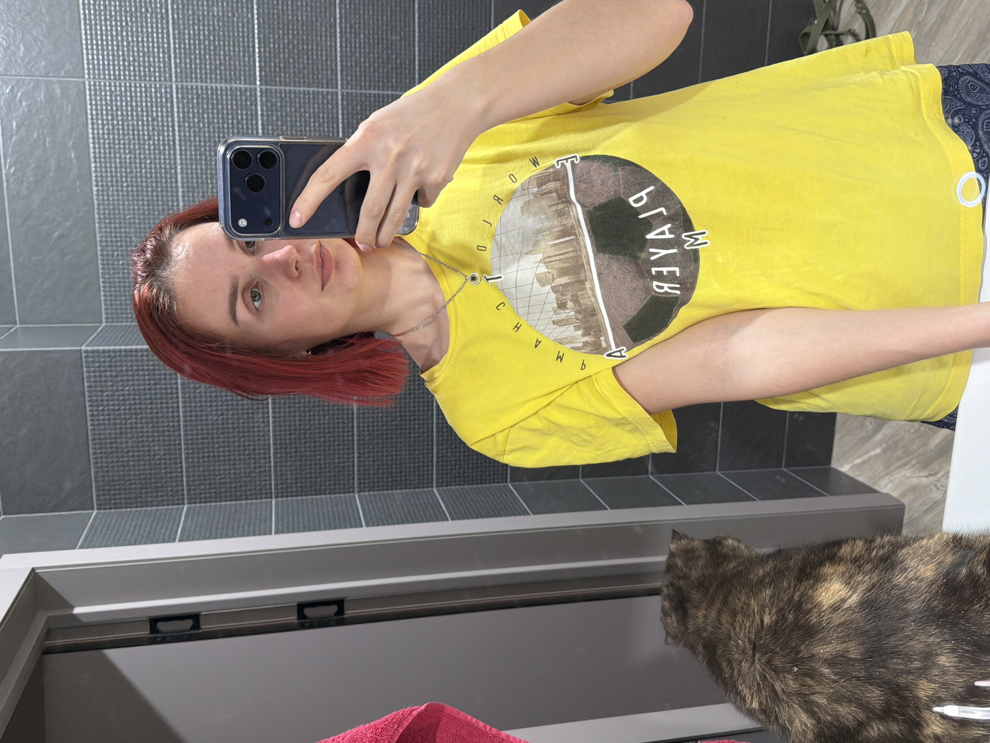 A woman with red hair takes a selfie in a bathroom with gray tiles. She is wearing a yellow t-shirt with a graphic print. The bathroom vanity is visible with a gray countertop. A cat is also in the frame. The location is a bathroom in a residential building in the city of Chaikovsky, Russia.
