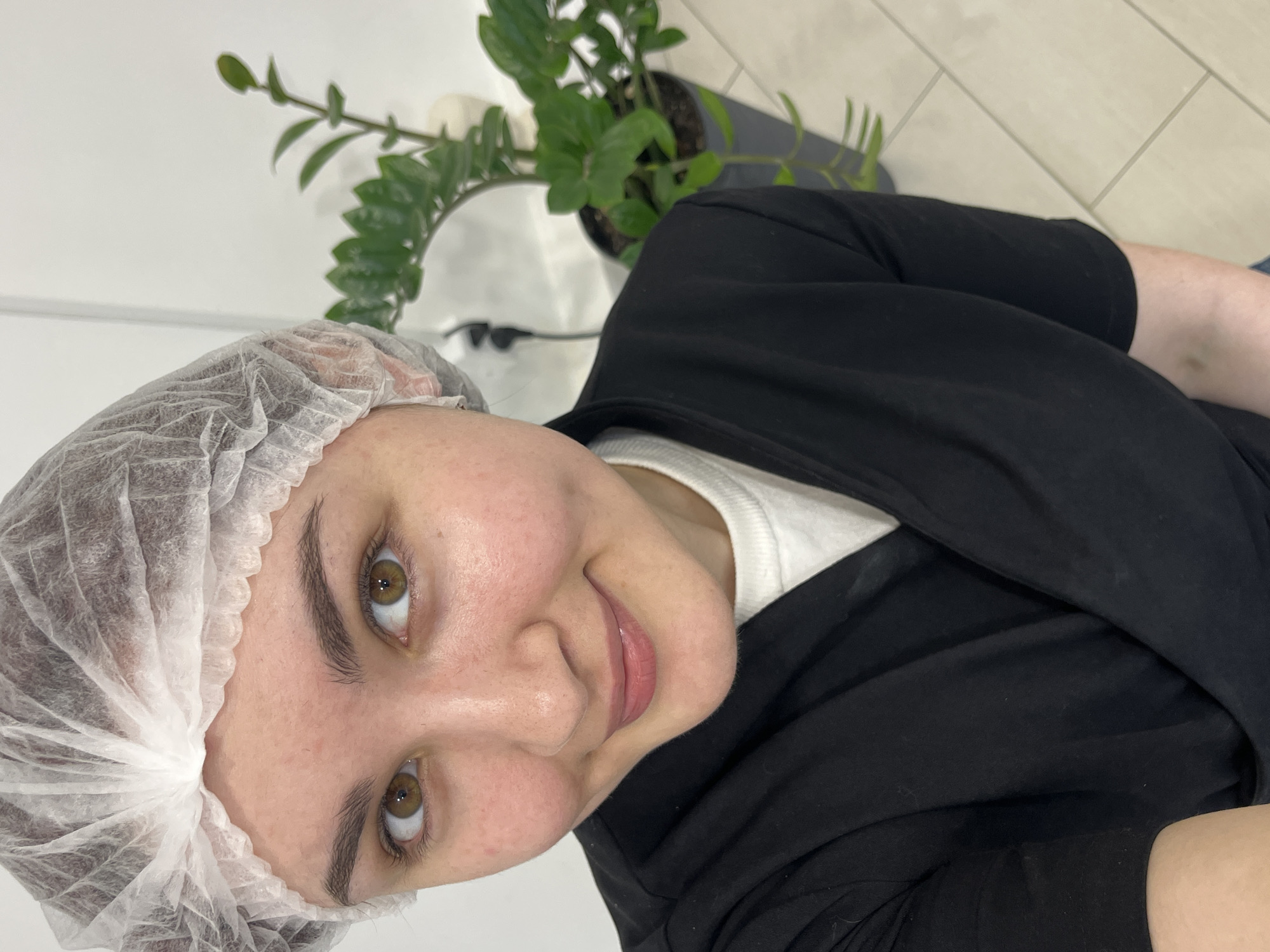 A person with fair skin, wearing a white disposable hairnet, is looking at the camera with a slight smile. They have brown eyes and are wearing a white shirt or undershirt beneath a black garment. A potted green plant is visible in the upper portion of the image behind the person. The background consists of a white wall and tiled floor. The image appears to be taken indoors. The location is provided as Neftekamsk, Russia.