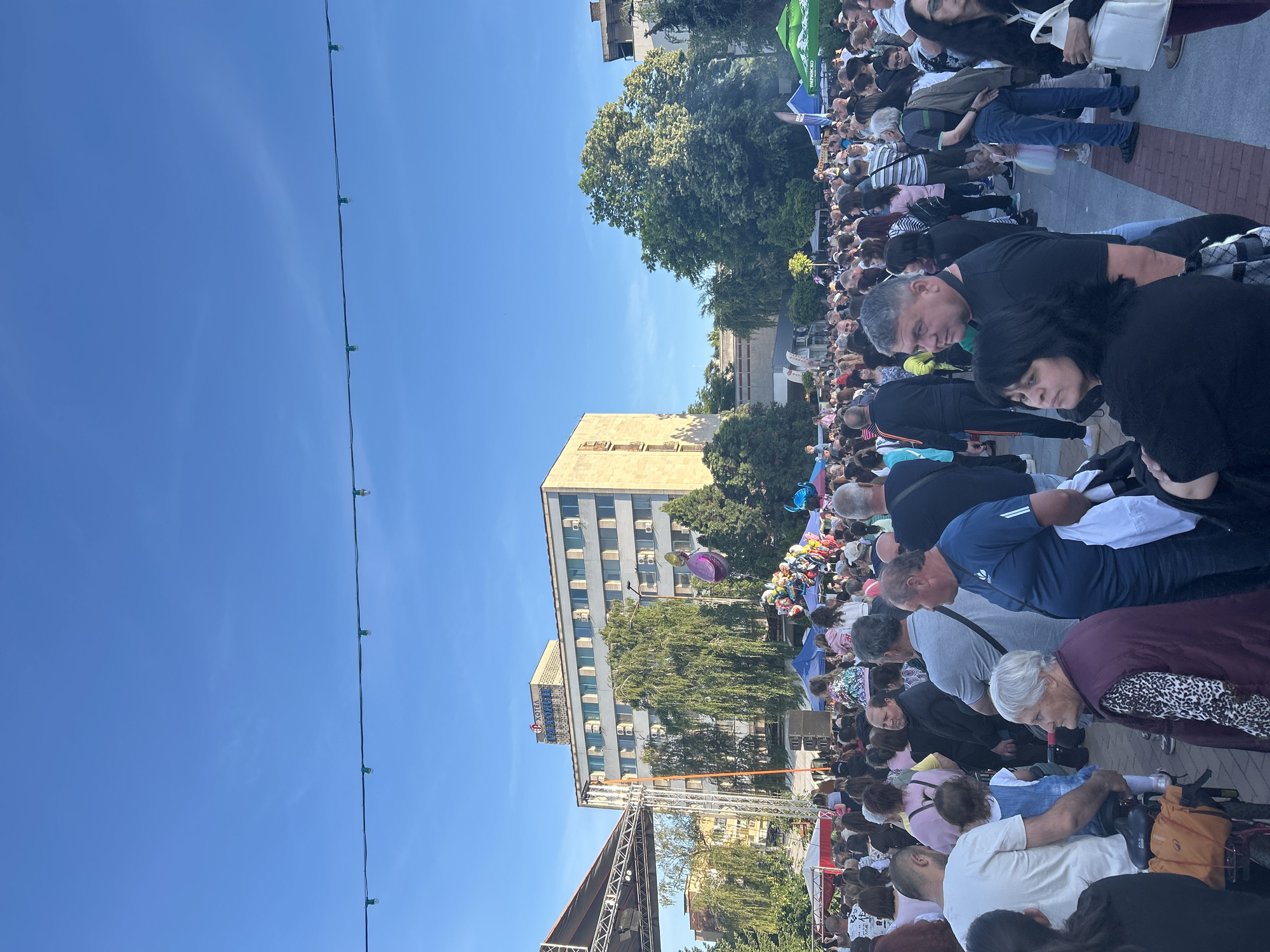 A crowd of people gathers in Gorna Oryahovitsa, Bulgaria. A stage is set up with a scaffold and a building stands in the background with the name "HOTEL" visible. A string of lights stretches across the sky. The sky is blue and the trees are green.