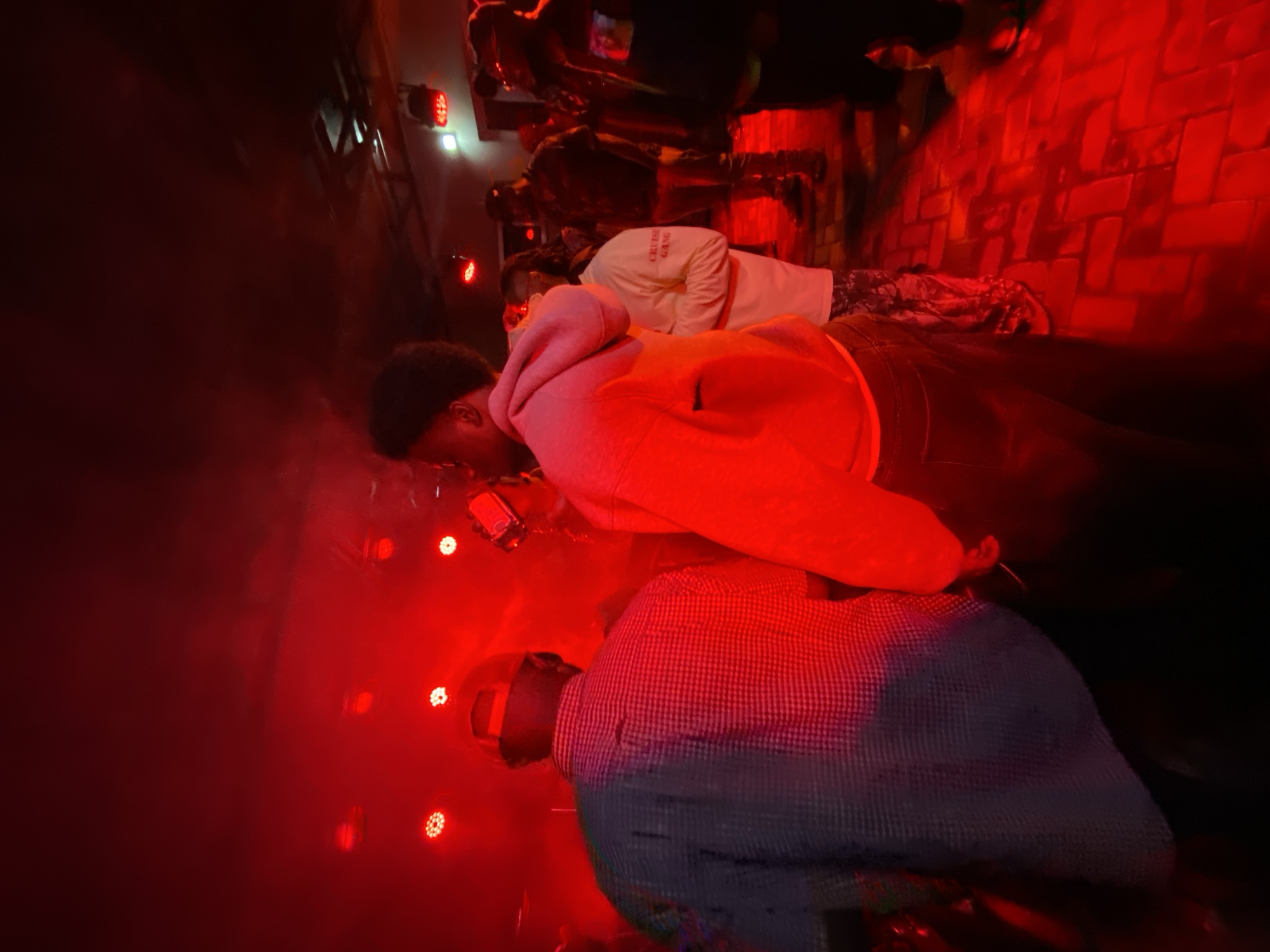 The image depicts an indoor scene illuminated by red stage lights, suggesting a concert or performance venue. Several individuals are present, with a focus on two men in the foreground. The man on the left is seen from behind, wearing a blue and white checkered shirt and a beige baseball cap. To his right, another man is partially visible, wearing a bright pink hoodie and dark pants. In the background, a crowd of people can be seen, some wearing casual attire. The environment appears to be a dimly lit room with a brick-patterned floor, also lit by the red stage lights. The atmosphere is energetic, consistent with a public gathering or event. The provided context indicates this scene is located in Abuja, Nigeria.