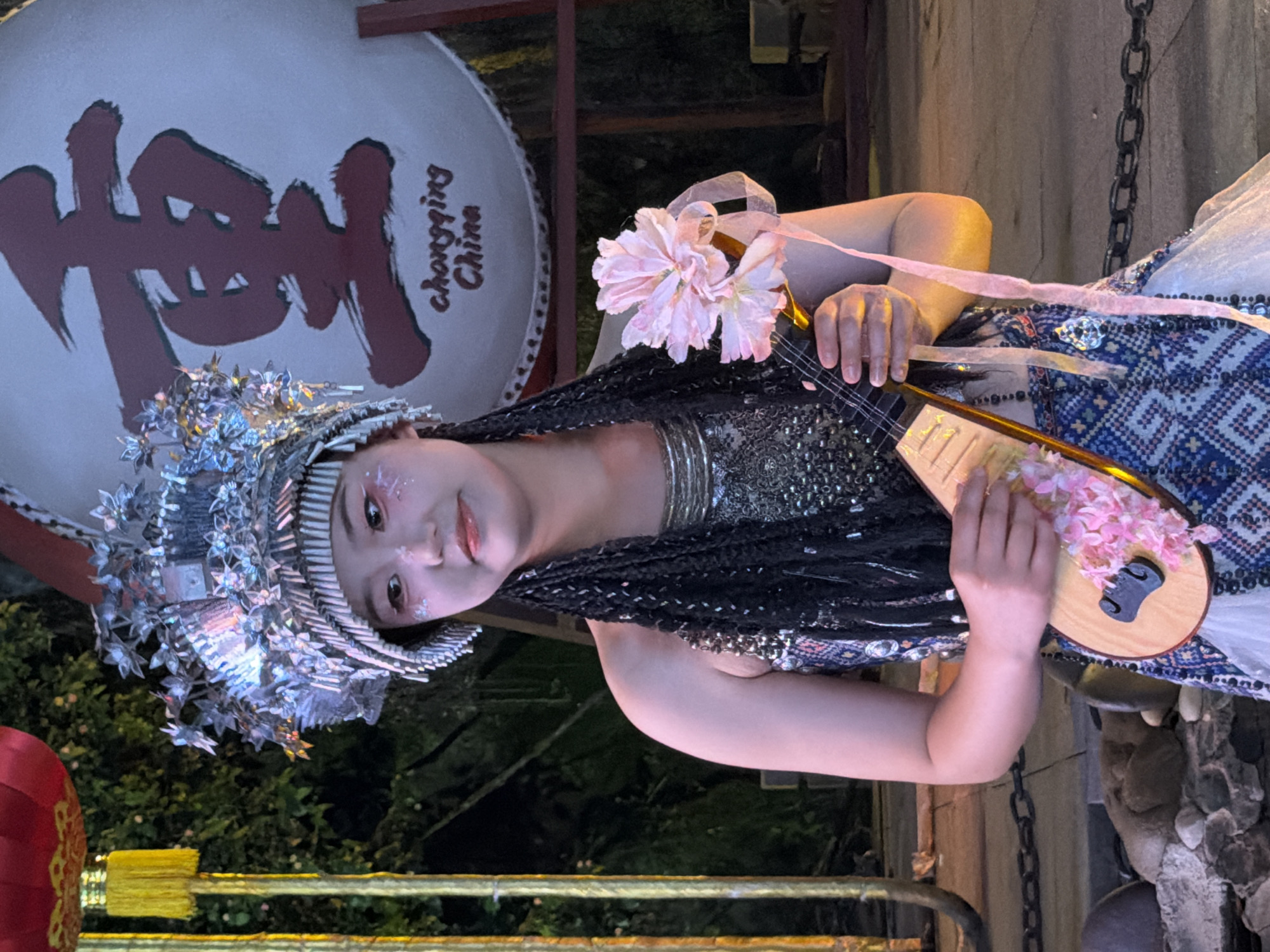 A young woman in traditional Chinese attire is the central subject, holding a pipa-like instrument decorated with pink flowers. She wears an elaborate silver headdress adorned with floral and crystalline elements, and her makeup features glitter on her eyes and cheeks. Her hair is styled in long, dark braids, intertwined with sparkles. She is dressed in a shimmering, possibly sequined, top and a patterned garment with blue and white geometric designs, complemented by a pink ribbon.
She is positioned in what appears to be an outdoor performance setting at night. Behind her, a large, round drum with a red, calligraphic inscription is visible, along with the text "Chongqing China". The background includes foliage and a rustic wooden structure with a metal chain. Red lanterns and a yellow railing are also present, suggesting a festive or cultural event. The lighting appears to be artificial, casting a warm glow on the performer and creating a dramatic ambiance. The woman has a gentle, perhaps serene, expression. The scene evokes a cultural performance or celebration, likely in Chongqing, China, given the visible text.