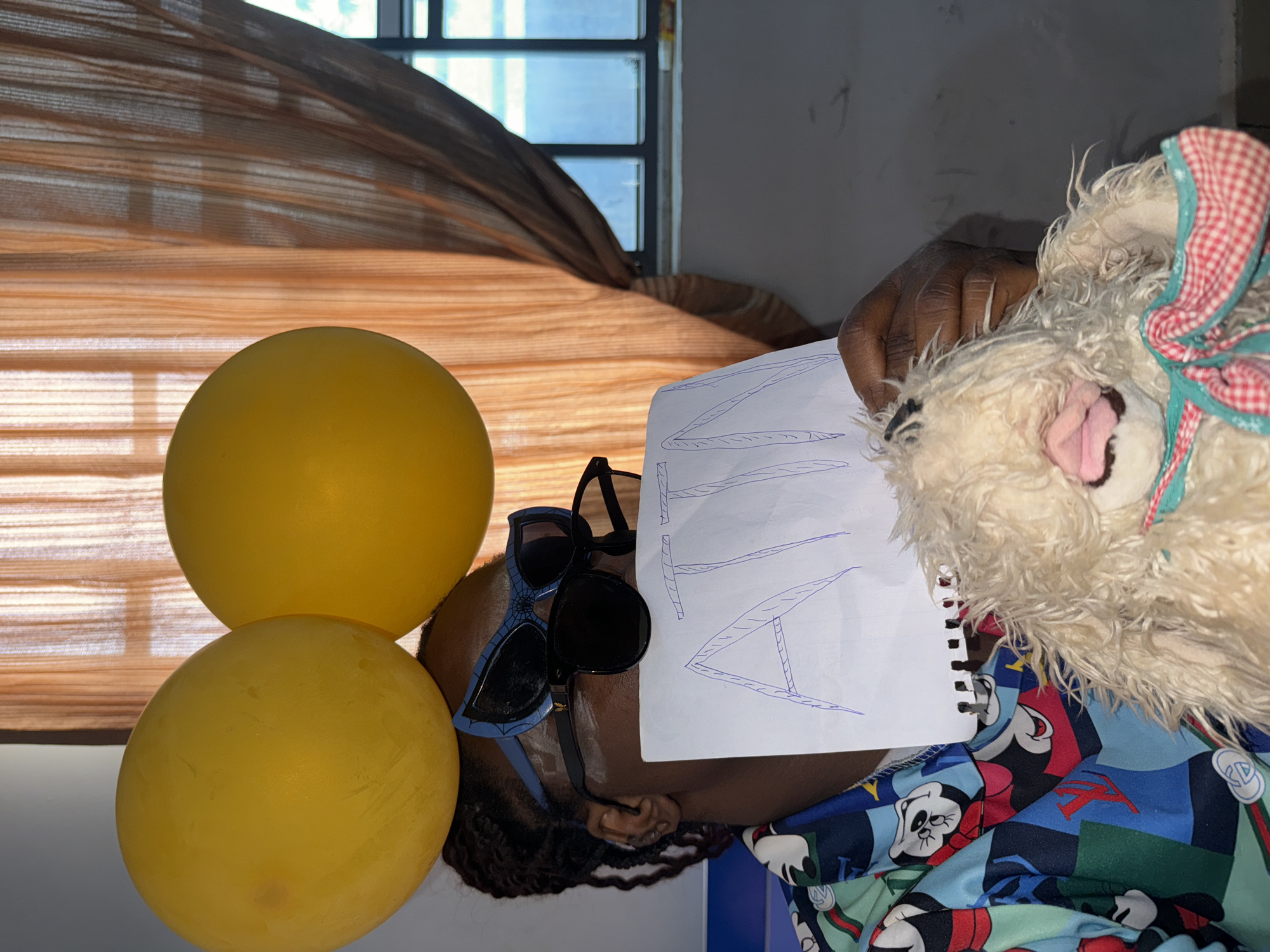 The image depicts a person indoors in Sabon Tasha, Nigeria. The person is wearing sunglasses, and two yellow balloons are positioned near their head. They are holding a piece of paper with writing in blue ink and a stuffed animal. The person is wearing a colorful patterned shirt. A window with closed blinds is in the background.