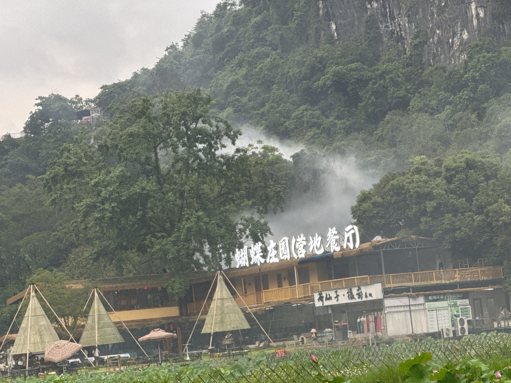 The image depicts a lakeside scene in Guilin, China. The foreground features a body of water covered with lotus plants, partially obscured by a wire fence. Behind the water is a two-story building, likely a restaurant or resort, adorned with a sign reading "蝴蝶庄园(营地餐厅)" ("Butterfly Manor (Camp Restaurant)"). Additional signage below the first sign reads "荷仙子、摄萄会社" ("Lotus Fairy, Photography Society"). The building has a wooden structure with an exposed balcony. In front of the building are tent-like structures with thatched roofs, arranged near tables and chairs indicating an outdoor dining area. A smaller building, likely a shop, stands to the right of the main building, next to air conditioning units. The background is dominated by a forested hillside, partially shrouded in mist or smoke. At the top of the hill, a structure resembling a small building or gazebo can be seen. The overall scene is overcast and foggy.
