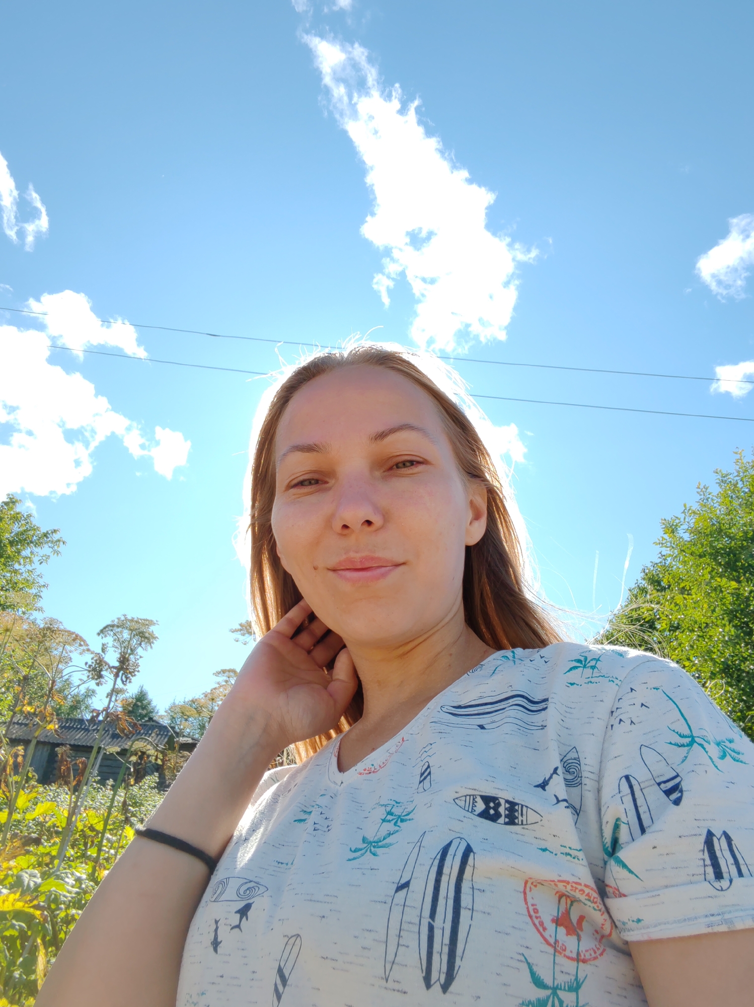 A young woman is pictured in an outdoor setting, with a bright blue sky and white clouds overhead. She is wearing a light-colored t-shirt with a pattern of surfboards and palm trees. Her right hand is raised to her neck. In the background, there are trees and vegetation, with what appears to be a wooden structure visible to the left. The lighting suggests it is daytime, with the sun positioned behind the woman, creating a slight backlight effect. The image is a selfie, taken from a low angle. The specific location of Чухлэм, Russia is not discernible from the visual cues in the image itself.