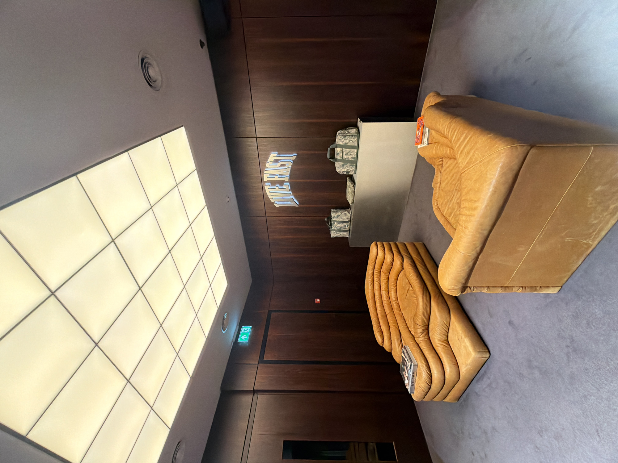 The image captures the interior of a building, potentially in London, United Kingdom. It features a dark wood-paneled wall with inset lighting and what appears to be a sign or logo that reads "EAST London". There's also an illuminated exit sign. Against the wall is a reception desk with luggage visible behind it. Two distinct, tan-colored, upholstered seating arrangements are present: one is a blocky, angular sofa, and the other has a ridged, organic shape. The floor is covered with a gray carpet. The overall lighting is a mix of artificial light from the ceiling panels and natural light sources.