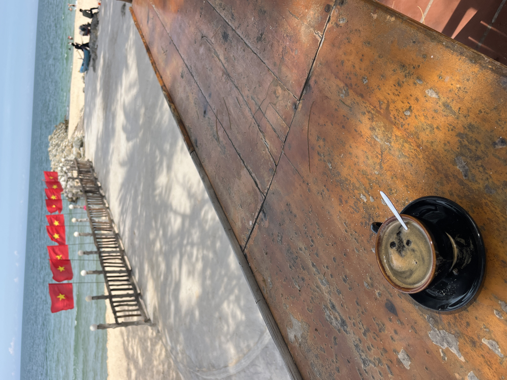 In Mũi Né, Vietnam, a rustic, weathered wooden table occupies the foreground, its surface showing signs of age and wear. On the table sits a dark brown ceramic cup of coffee, placed on a matching black saucer. A white spoon rests inside the coffee, with visible coffee grounds floating on the surface. In the background, a sandy beach stretches out towards the ocean. A wooden fence, adorned with several red flags bearing a yellow star (the flag of Vietnam), lines the edge of the beach. Beyond the fence, the turquoise ocean meets a clear blue sky. Further down the beach, a small blue boat is pulled ashore, with a few silhouetted figures visible near it, suggesting a few people are present on the beach. The scene captures a relaxed, coastal atmosphere with a patriotic touch.
