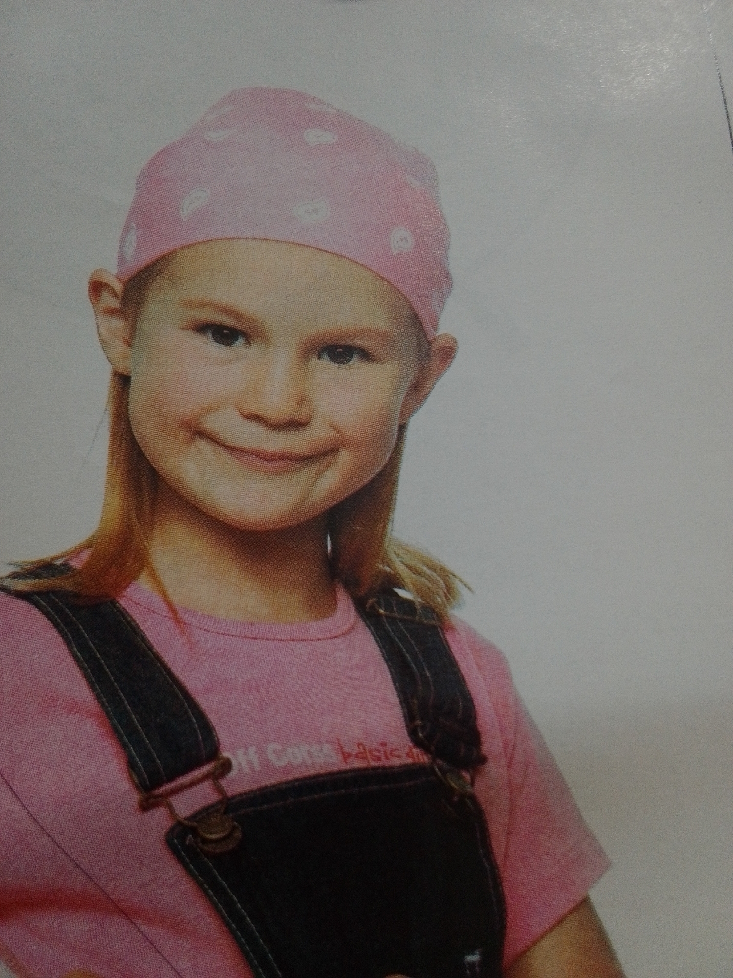 A young girl with blonde hair, wearing a pink bandana and a pink t-shirt with dark overalls, smiles at the camera. The t-shirt has white text that appears to read "Off Corses Pasic...". The girl is positioned in the center of the frame, with her face slightly turned to the left. Her expression is happy and she has her eyes open, looking directly at the viewer. The background is a plain, light color, suggesting a studio setting or a neutral backdrop. There are no obvious indications of time of day, weather, or specific location cues within the image itself.