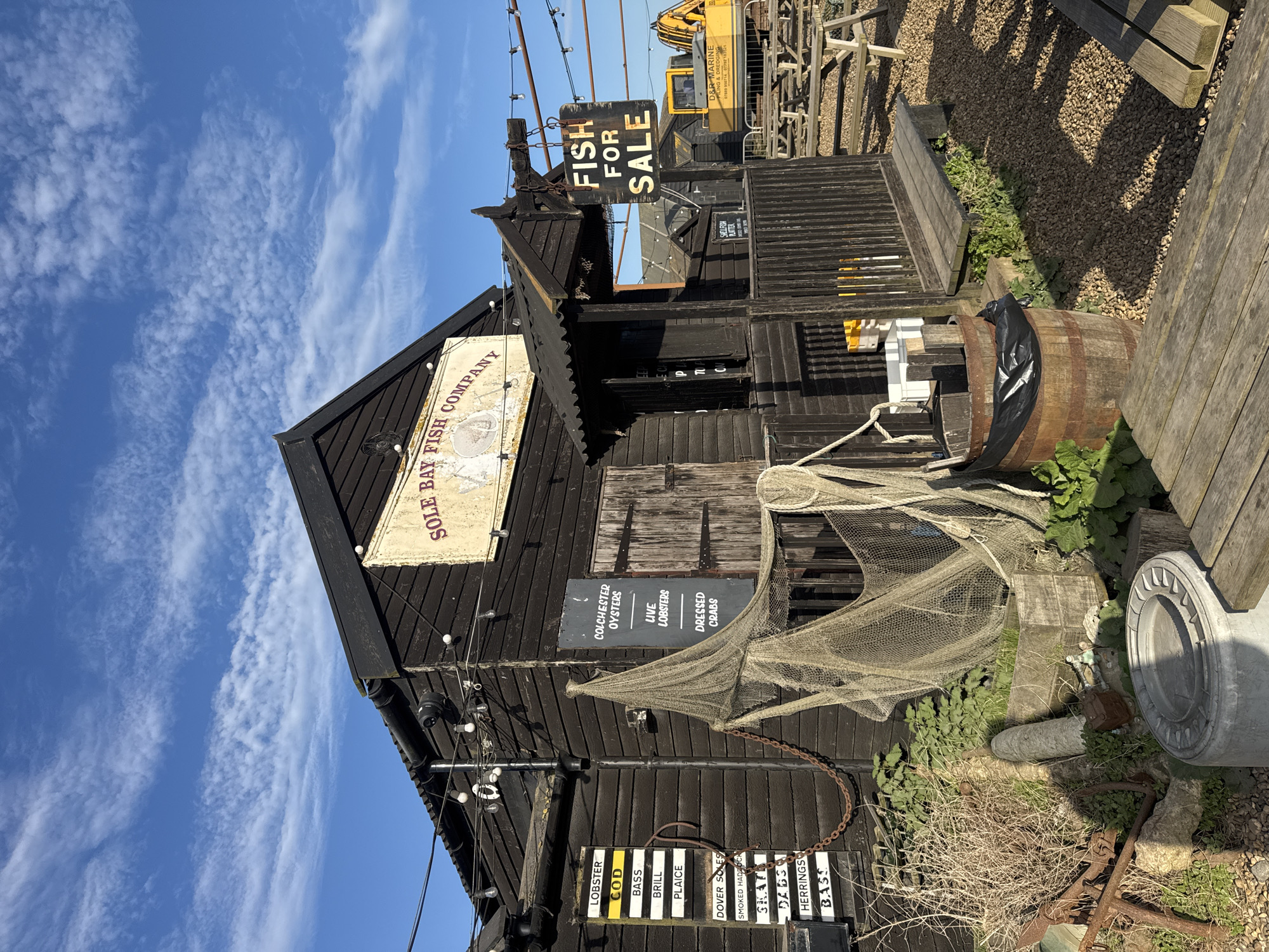 The image shows a fish market building in East Suffolk, United Kingdom, named "SOLE BAY FISH COMPANY". A prominent sign declares "FISH FOR SALE." The building has dark wood siding and various signs listing available seafood, including lobster, cod, bass, brill, plaice, Dover soles, smoked haddock, skate, dabs, herrings, and bass. Another sign advertises "COLCHESTER OYSTERS, LIVE LOBSTERS, DRESSED CRABS." A fishing net is draped nearby, adding to the maritime atmosphere. Additional debris is scattered around the bottom, including a wheel and chain. A barrel sits to the right of the front door. A yellow construction vehicle is in the background.