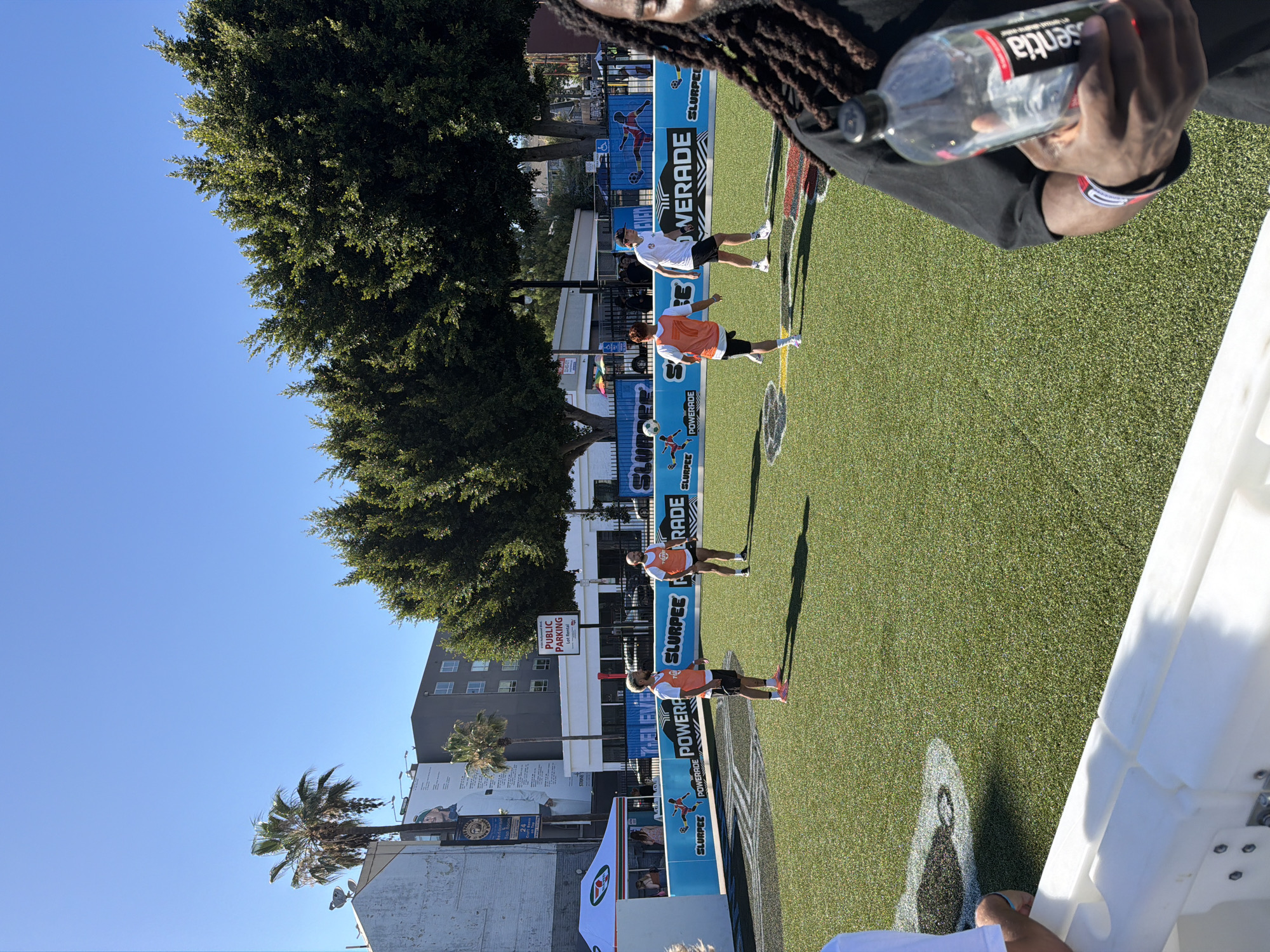 A group of people are playing soccer on a bright, sunny day in Los Angeles, California. The soccer field is artificial turf, and the surrounding area is paved with a white barrier. There are buildings in the background, including a tall building with a billboard. One person in the foreground is holding a bottle of water. There is a large banner in the background with the words "SLURPEE" and "POWERADE" on it. The sky is clear and blue.