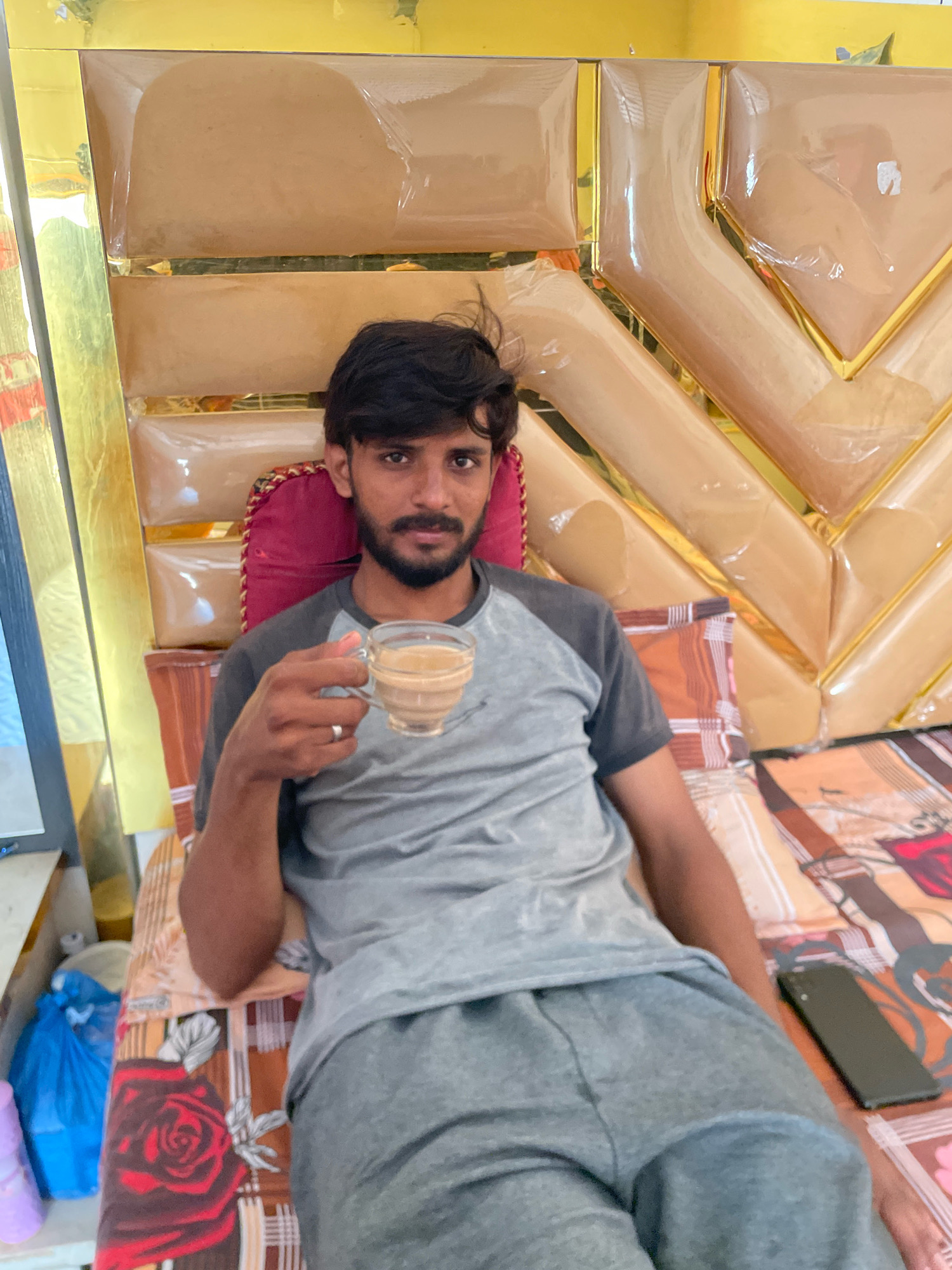 A young man with dark hair and a beard is seated on a bed with a patterned comforter, holding a glass of tea. He is wearing a gray t-shirt with dark gray sleeves and gray sweatpants. The bed has a distinctive headboard with padded, diamond-shaped sections in a light tan color, outlined in gold trim. The background includes a wall with gold paneling. To the left of the man, partially visible, is a television screen. On the comforter to the right of the man, a dark-colored smartphone is placed. In the lower left portion of the image, there are two plastic bags, one blue and one lighter-colored, along with a light purple cylindrical object, possibly a bottle. The scene appears to be an indoor setting. While the image is not explicitly dated, the style of the furniture and the overall setting suggest a contemporary domestic environment. The location is specified as Chak 90 Morr, Pakistan.