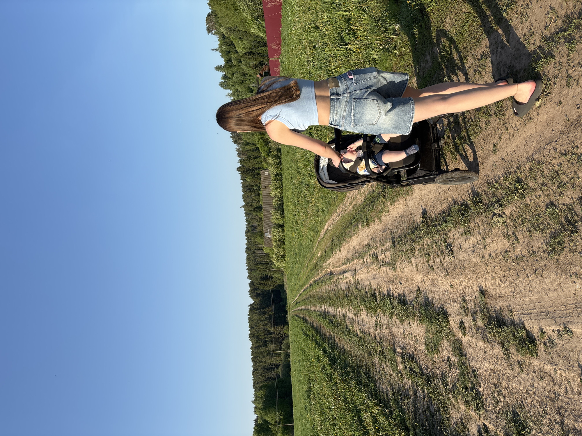 A woman stands beside a baby stroller on a dirt path in Lukintsy, Russia. The stroller, which contains a baby, is black with small wheels. The woman has long brown hair, and she wears a light blue top and denim shorts. She has bare legs and wears dark shoes. The dirt path is partially covered with grass, and the vegetation on either side of the path appears to be sparse. A treeline is visible in the background. The sky is clear and blue.