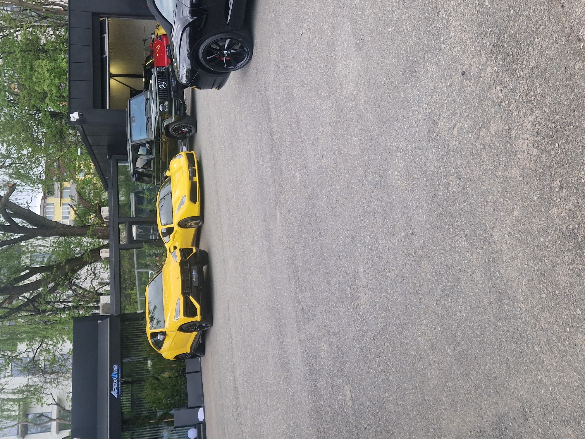 The image, rotated 90 degrees clockwise, captures a scene outside a modern building, likely a luxury car dealership or service center. The foreground features an asphalt pavement.
The primary subjects are several high-end vehicles. Two bright yellow Lamborghini Urus SUVs are prominently parked in the foreground, facing towards the right. Behind them, closer to the building, a dark green Mercedes-Benz G-Wagen is parked. A black car, possibly another SUV or a large sedan, is partially visible on the far right, with its front wheel showing. Inside the building's garage opening, a red car is also partially visible.
The building itself is sleek and dark-colored, possibly black, with large windows or open bays. On the side of the building, a prominent white sign with blue accents reads "Apex One LUXURY CARS". The text "ApexOne" is also visible on the front grille of the yellow Lamborghini Urus closest to the viewer.
The setting appears to be an urban or suburban environment. Mature trees with green foliage are visible to the left and in the background, along with other residential-style buildings with multiple windows. The lighting suggests it is daytime, possibly overcast, as there are no harsh shadows. No people are visible in the frame.