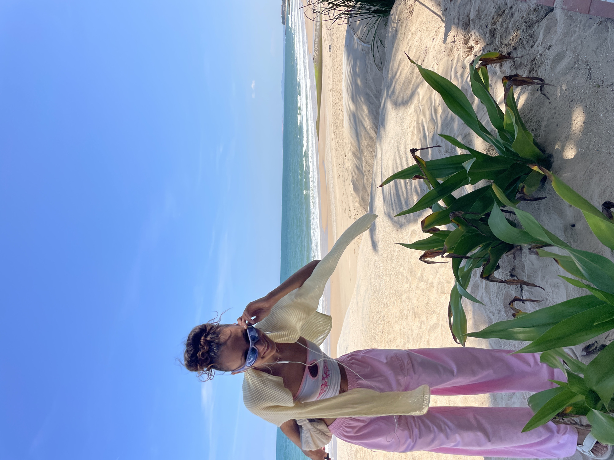 A young woman stands on a sandy beach in Phu Thuy Ward, Vietnam, adjusting her sunglasses. She has her hair pulled up in a bun and is wearing a white top, a light yellow cardigan draped over her shoulders, and pink sweatpants. She is also wearing white earbuds and white sandals. In front of her, green plants with some brown, dried leaves are growing in the sand. The ocean is visible in the background, with waves gently rolling onto the shore under a clear blue sky. The beach appears to be a public area, with the sand shaped by wind and possibly footprints.