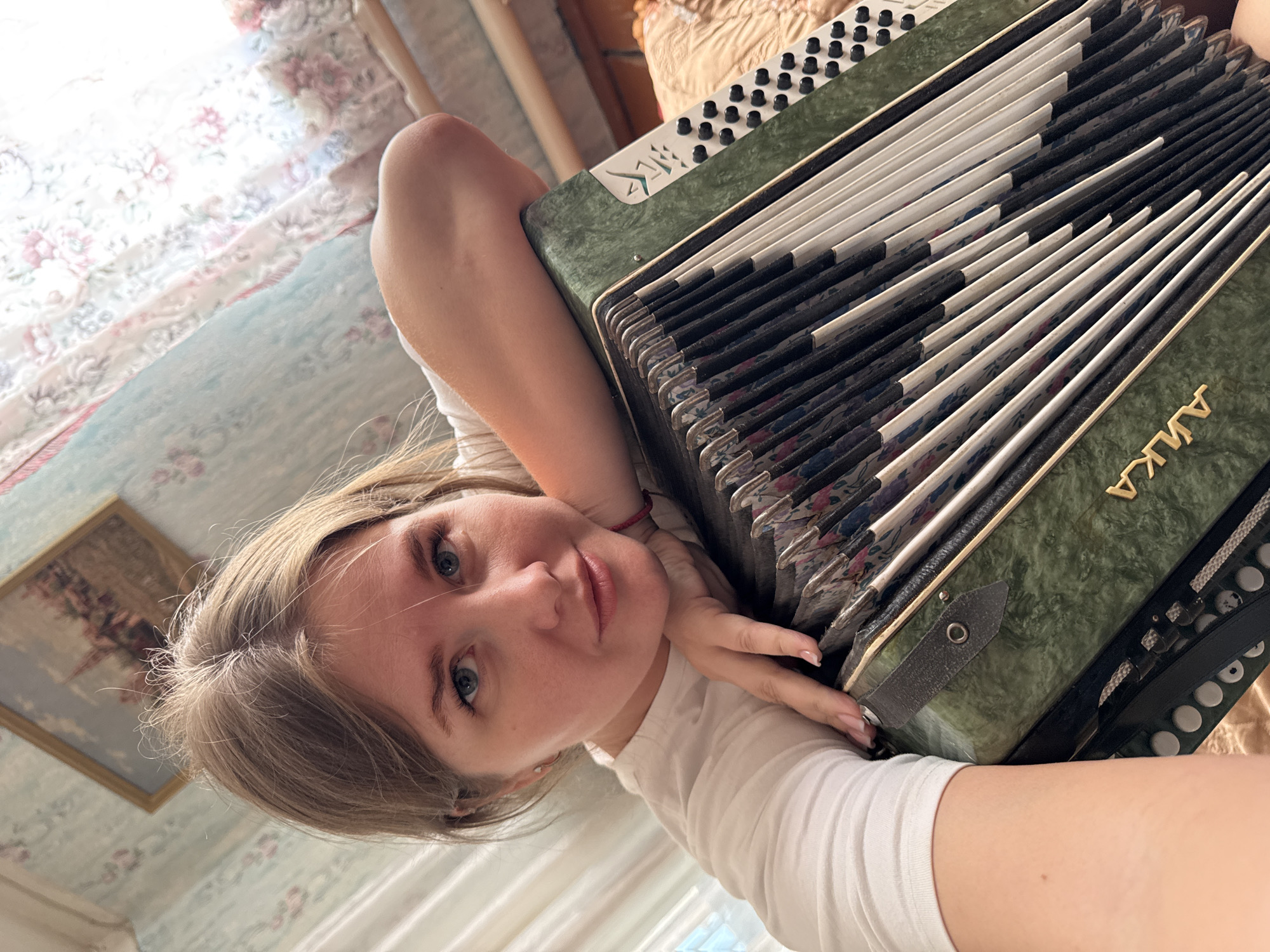 A woman lies on a patterned blue and white bedspread, leaning against a large green accordion. She wears a white t-shirt, has fair skin, and blonde hair. The accordion’s keys are white and black with a floral patterned bellows. The brand name “ANKA” is visible on the accordion. A framed picture hangs on a wall in the background. The location appears to be indoors, potentially a home. There is no identifiable evidence to confirm the location as Фоки, Russia.