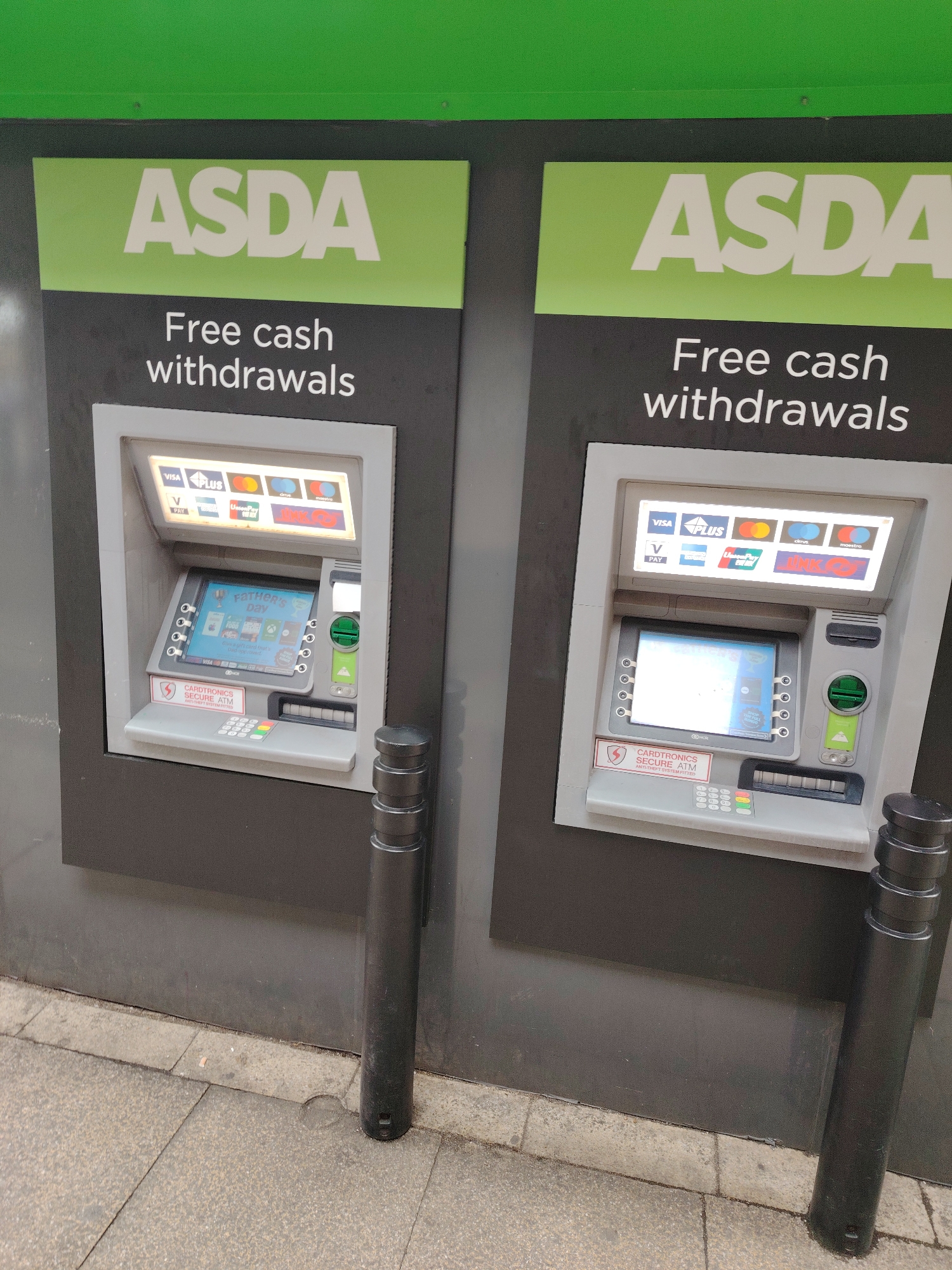 This image displays two side-by-side automated teller machines (ATMs) located outdoors at an ASDA facility in Peterborough, United Kingdom. The ATMs are installed into a dark grey wall, which features a bright green fascia board at the top. Above each ATM, a light green panel prominently displays the word "ASDA" in large white capital letters, with "Free cash withdrawals" written below it in white text on a dark grey background.
Both ATMs are identical Cardtronics Secure ATM models, equipped with anti-theft systems as indicated by a red and white label on the lower left of each machine's console. An illuminated panel above the main screen on each ATM displays logos for accepted card types, including Visa, Plus, V PAY, UnionPay, Cirrus, and Maestro. The central digital screens of both ATMs are active, currently displaying a promotional graphic related to "FATHER'S DAY," featuring various gift and food imagery. Each ATM includes a standard numerical keypad with distinct green (enter), yellow (clear), and red (cancel) buttons, a card insertion slot below the screen, and a cash dispensing slot below the keypad. Additional control buttons are arranged vertically along both sides of the screen.
Two black, cylindrical bollards, featuring a stacked-ring design, are concreted into the grey, textured pavement directly in front of each ATM, likely serving as protective barriers. The pavement consists of worn concrete slabs, with a small piece of light-colored debris visible near the base of the wall between the bollards. The overall scene is devoid of people or any active transactions.