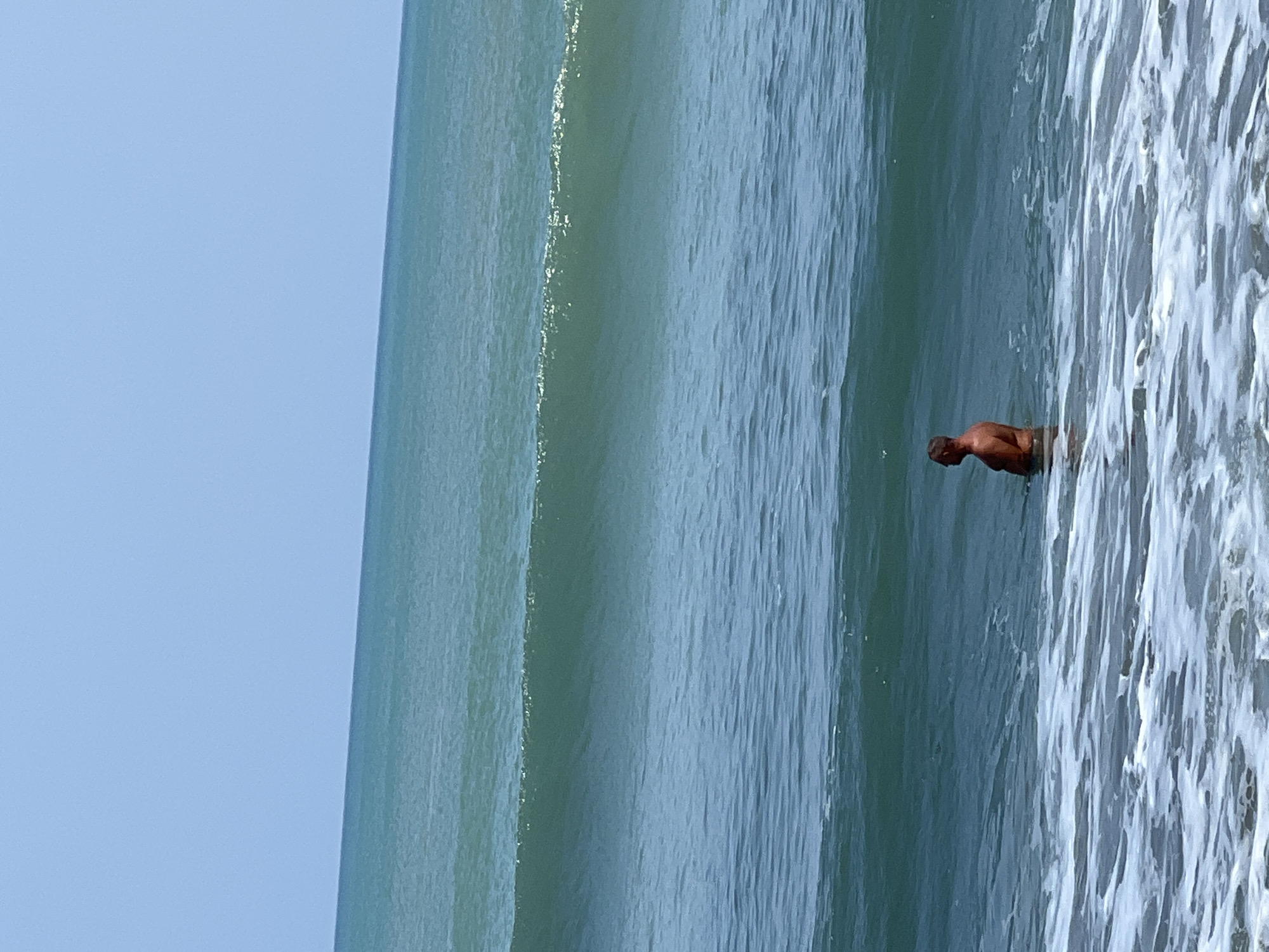 A man stands in the ocean in Kuta, Indonesia. The man is shirtless and appears to be waist-deep in the water. He is facing to the left, with his profile visible. He has short, dark hair and a muscular build. The ocean is a deep blue-green color, with several waves visible. The sky is clear and light blue. The image appears to have been taken from the shore, looking out at the ocean.