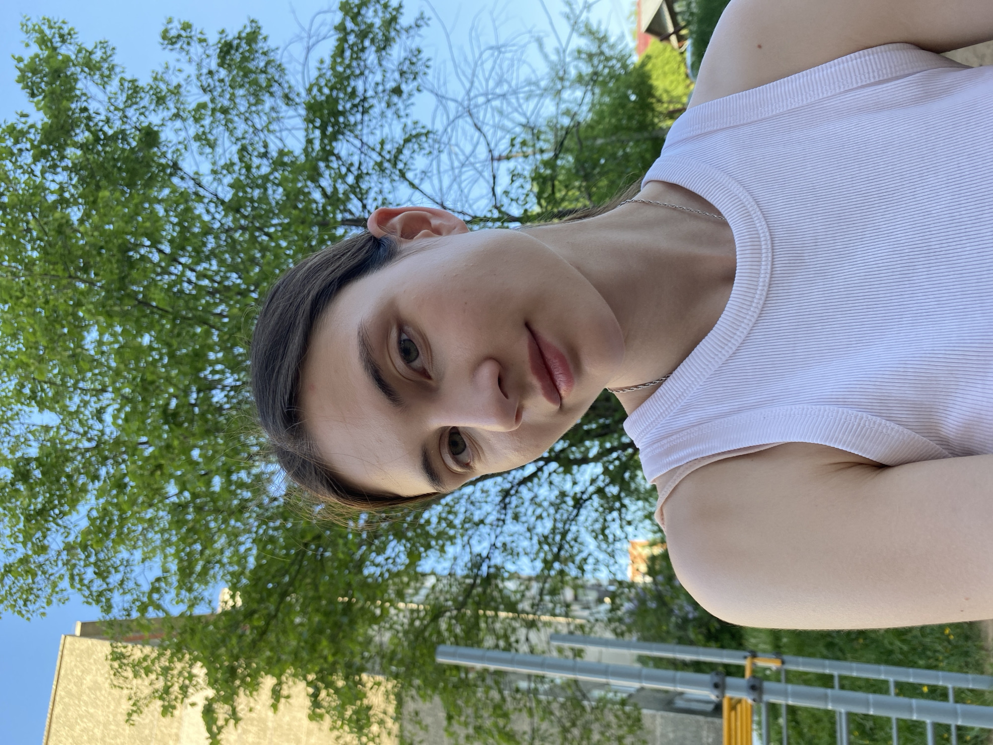 A young, fair-skinned woman with dark hair is seen from a low-angle perspective. She is wearing a white ribbed tank top and a silver necklace. The background includes green foliage, possibly trees, a building, and a blue sky. The woman appears to be outside in Votkinsk, Russia. The city environment includes building architecture and greenery typical of urban settings. The image captures a moment of direct eye contact from the subject. Construction scaffolding in the background.