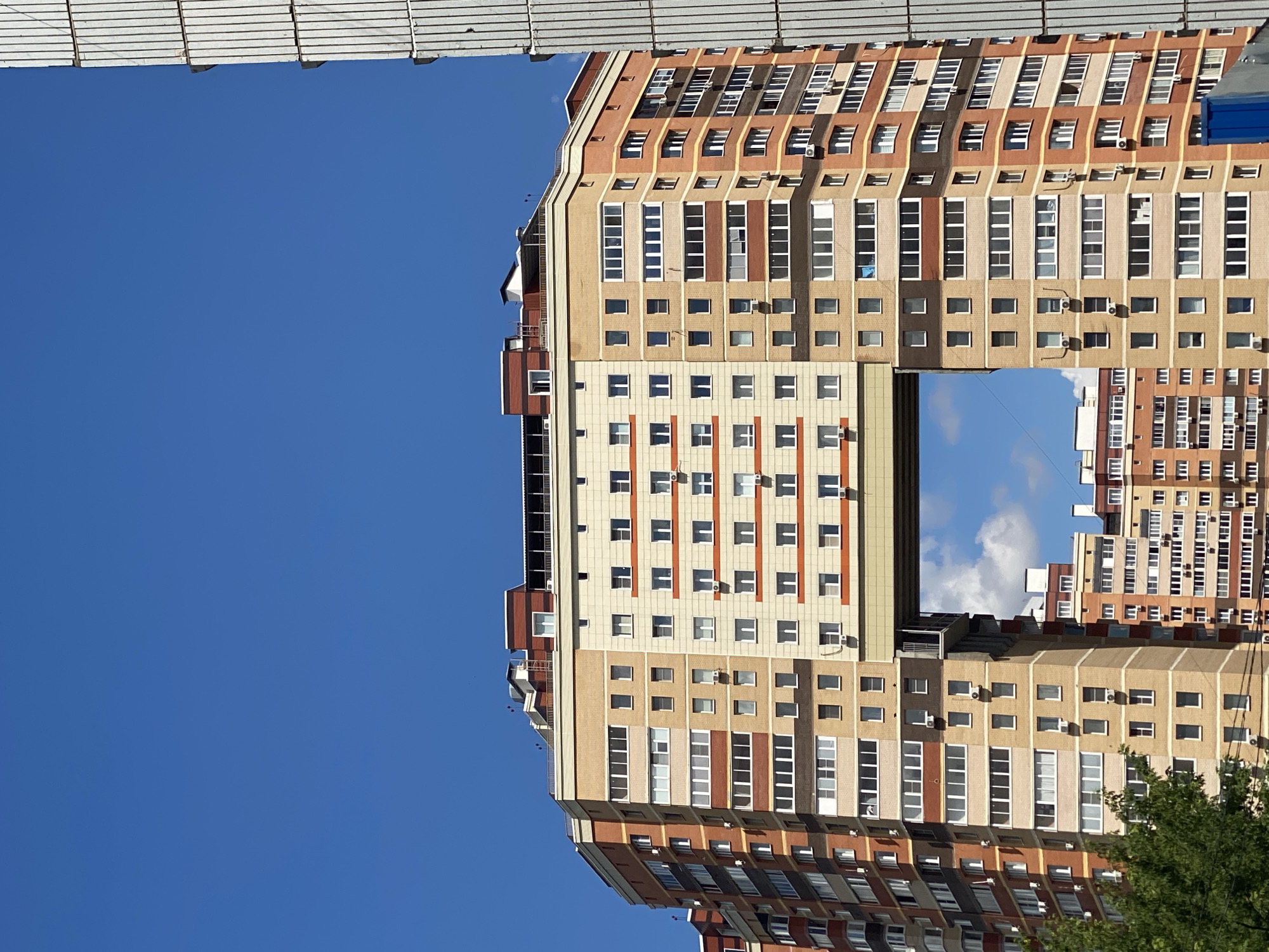 This is a low-angle, vertical shot of a modern apartment building in Kotelniki, Russia, under a clear blue sky. The building, primarily constructed with tan and brown brickwork, features numerous windows with white frames. Some windows are rectangular and grouped in vertical columns, while others are smaller and square, arranged in horizontal rows. Many of the square windows have air conditioning units mounted below them.
The building's architecture is characterized by stepped levels and a central open section that reveals a patch of sky with some clouds. The topmost section of the building has a distinctive wavy roofline. In the lower right corner, the green leaves of a tree are visible, suggesting the presence of vegetation in the vicinity. The overall impression is of a bright, sunny day. No people are visible, and the image appears to capture a static view of the residential structure. There is no discernible text in the image.
