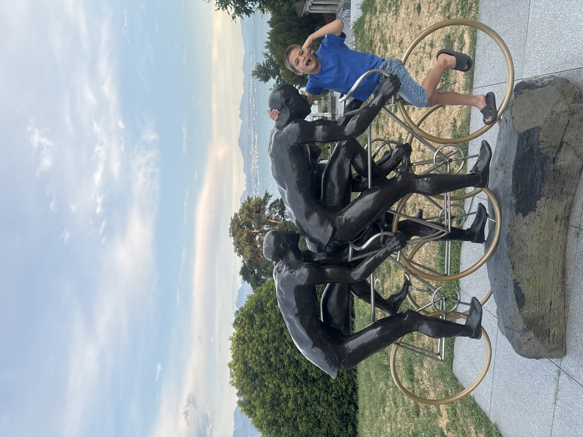 The image depicts a sculpture of three cyclists in motion, positioned in the Parc de Mon-Repos in Lausanne, Switzerland. A young boy, wearing a blue t-shirt and denim shorts, is interacting with the sculpture. He is standing on one of the bicycle wheels and has his hands near his face, appearing to be making a gesture. The sculpture itself is made of dark material and is mounted on a large rock. In the background, a body of water, likely Lake Geneva, is visible, with mountains and trees on the horizon. The sky is partly cloudy.