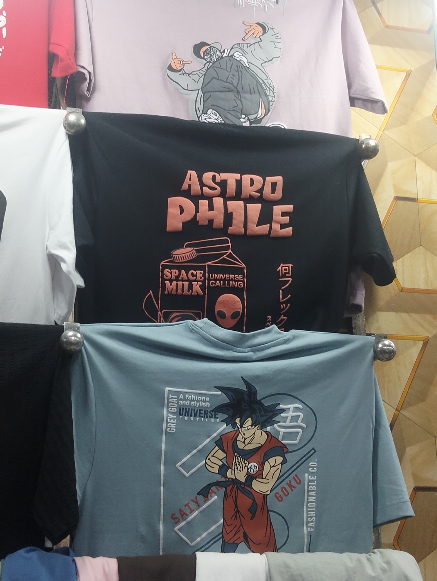 The image shows a rack of T-shirts displayed for sale, likely inside a shop in Dhaka, Bangladesh. The shirts feature various graphic designs. The top shirt is lavender and has a cartoonish illustration of a person gesturing with their hand. The middle shirt is black with the words "ASTRO PHILE" in raised peach-colored lettering above a stylized drawing of a milk carton labeled "SPACE MILK, UNIVERSE CALLING," including an alien face. The bottom shirt is light blue with a graphic of Goku, the main character from the anime series Dragon Ball Z, along with the word "GOKU" in bold font. The background consists of a wall with a yellow geometric pattern. The shirts are hung on a rack with silver ball-shaped ends.