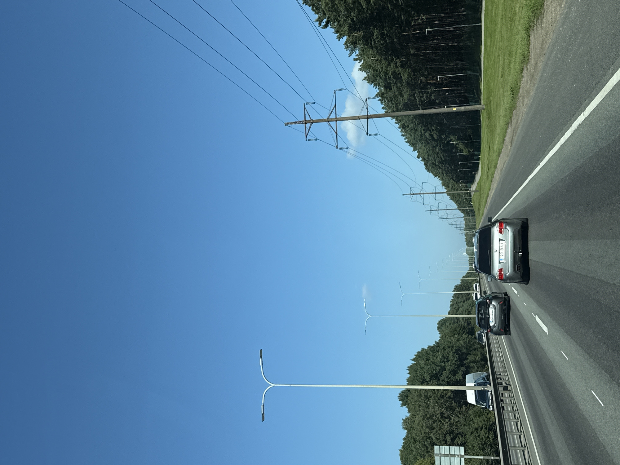 The image captures a view from a vehicle traveling on a multi-lane highway under a clear blue sky. To the left, a dense line of green trees borders the highway, with several modern streetlights spaced along the roadside. A white van is visible in the distance on the left. On the right side of the highway, a dense forest of tall pine trees stretches into the distance. High-voltage power lines supported by tall transmission towers run parallel to the road, also stretching into the distance. Several cars are on the highway, including a silver BMW in the foreground. The road surface is asphalt with visible lane markings. The image appears to be taken on a sunny day.